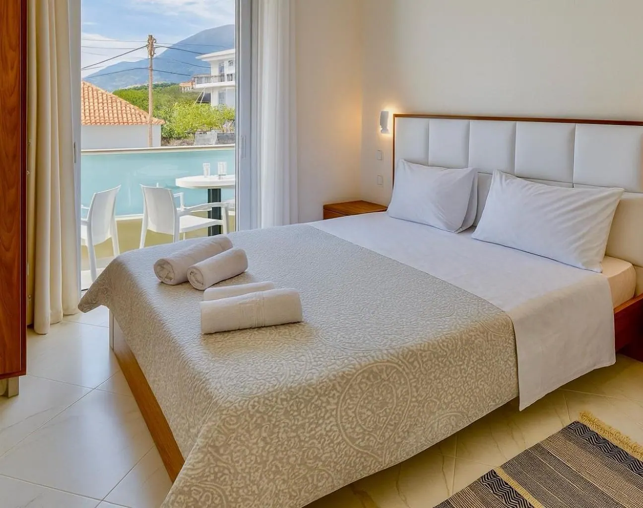 A double bed with a white padded headboard and walnut bedside tables, glass doors open to a balcony with the mountains beyond