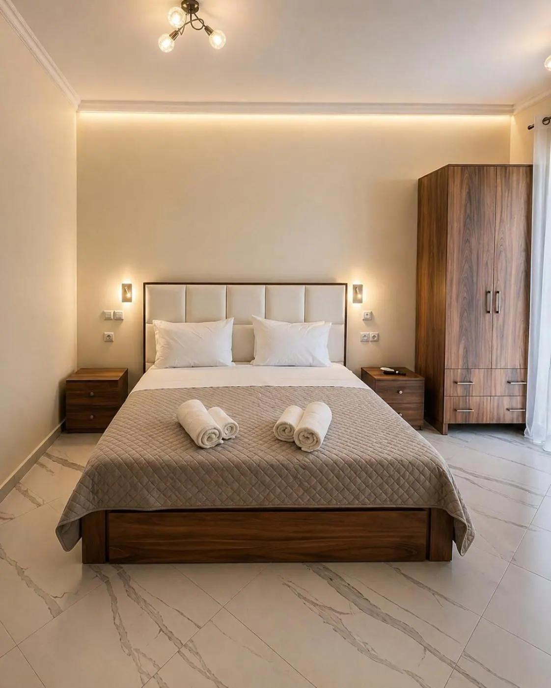 A double room in the evening, warm light along the ceiling cove, a padded headboard, walnut wardrobe and a marble-look floor