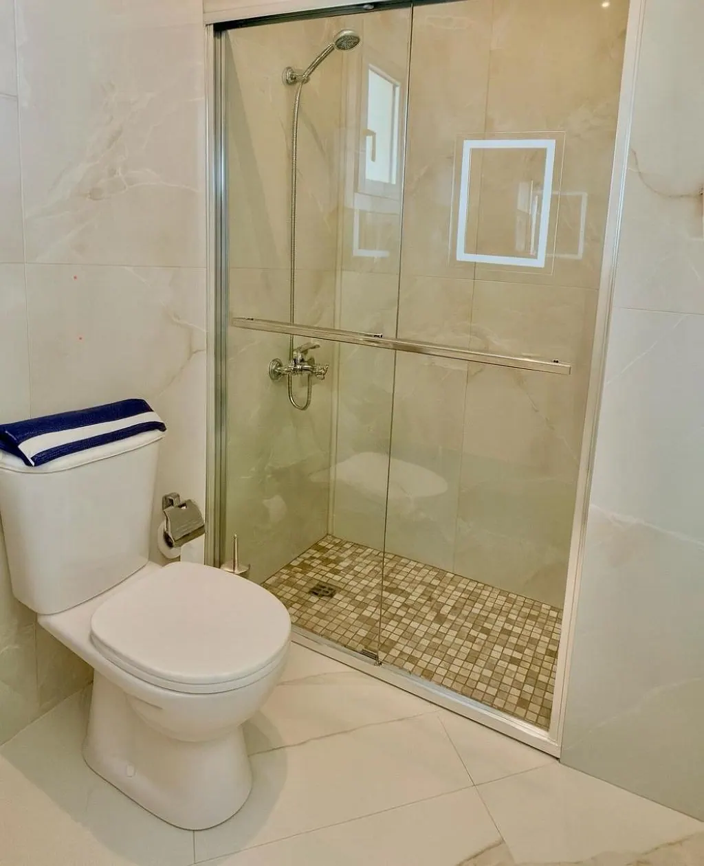 A shower room in cream marble-look tile with a glass shower enclosure, a pebble shower floor and a white toilet