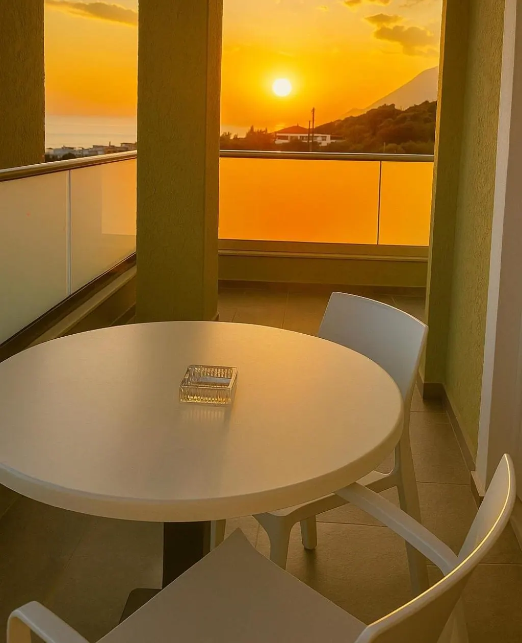 The sun setting over a headland, seen from a balcony with a round white table and white chairs, the whole balcony lit orange