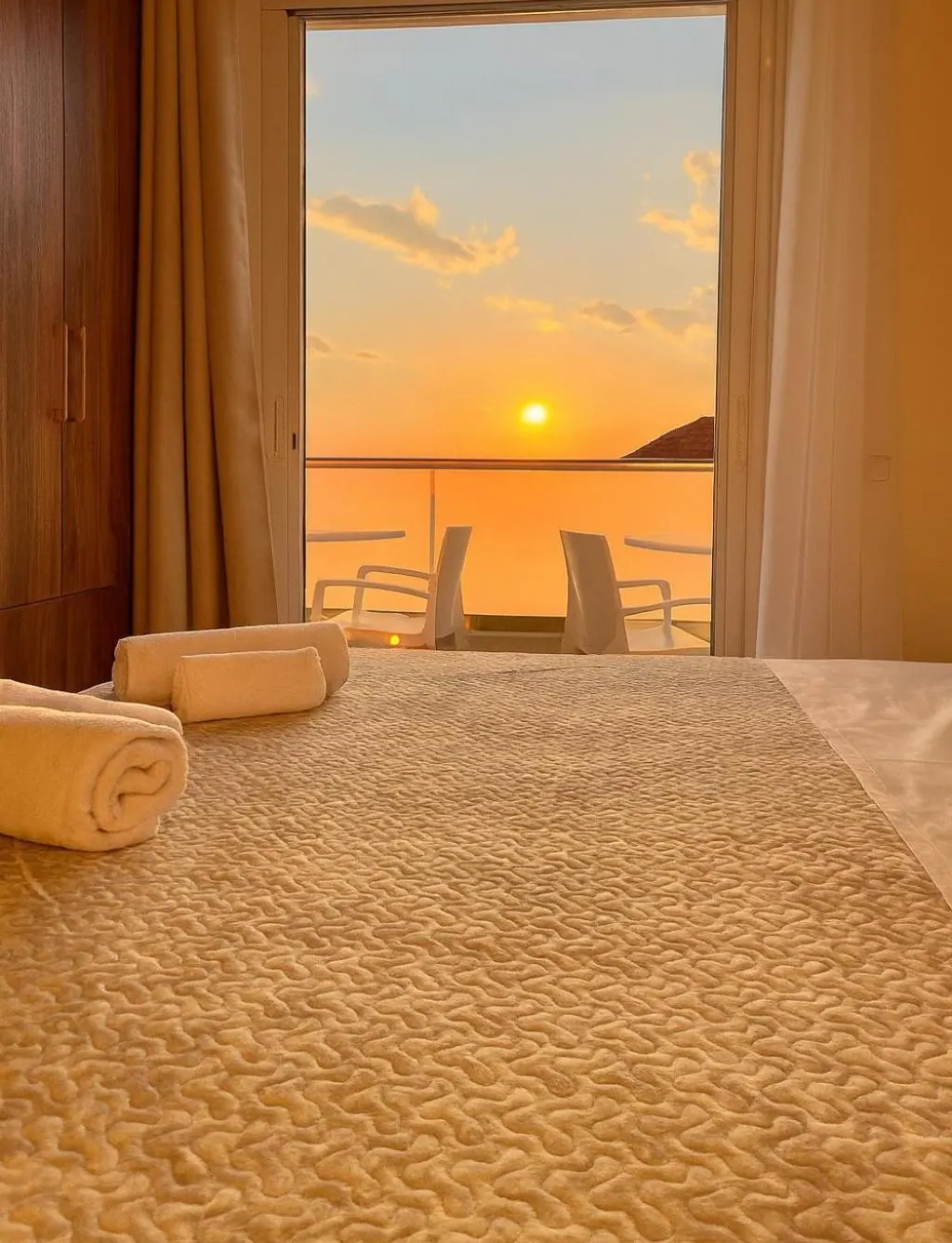 Rolled towels on a bed facing an open balcony door, the sun setting over the sea beyond the balustrade
