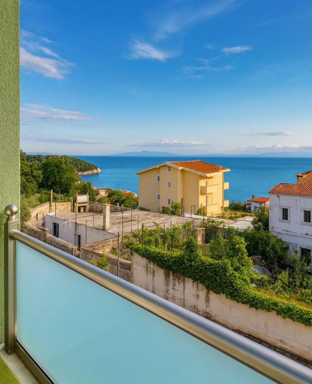 A balcony with a pale aqua glass balustrade looking down over olive gardens and red-roofed houses to the sea and a long island on the horizon