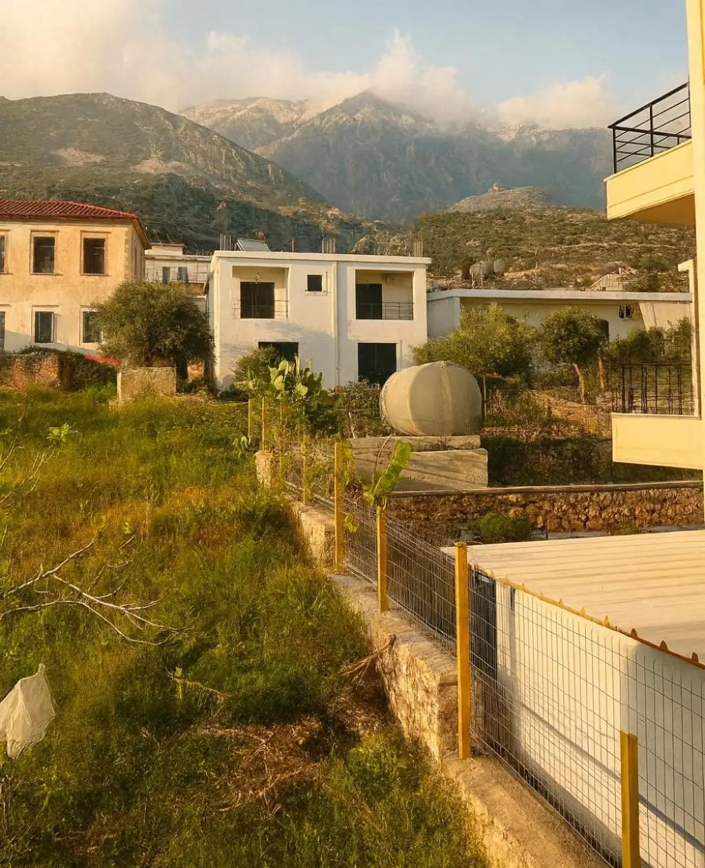 Wild grass and a dry-stone wall in front of two old village houses, with the mountains above them in late sun