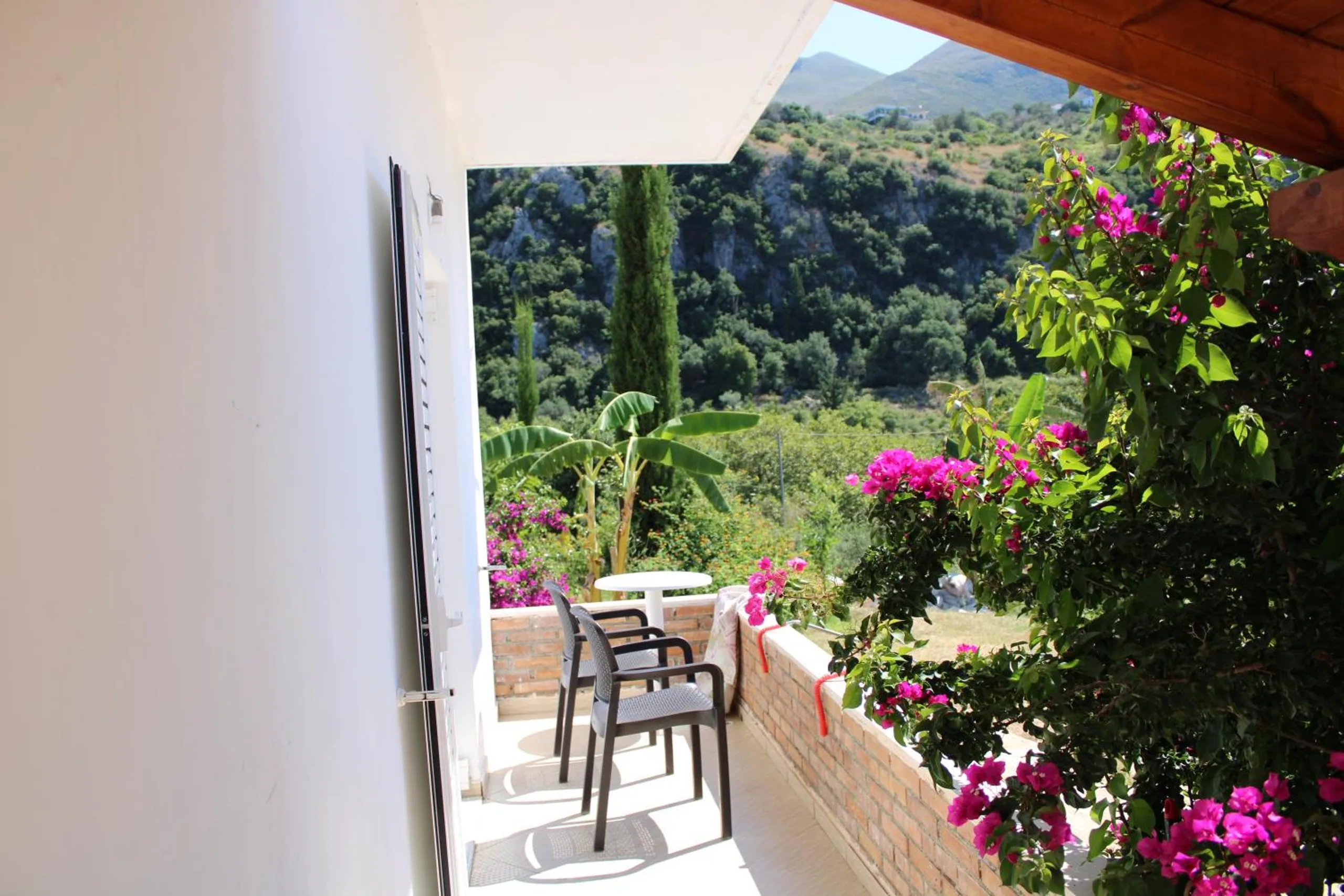 A balcony with chairs and a small table seen through an open door, bougainvillea on the rail and green hills beyond