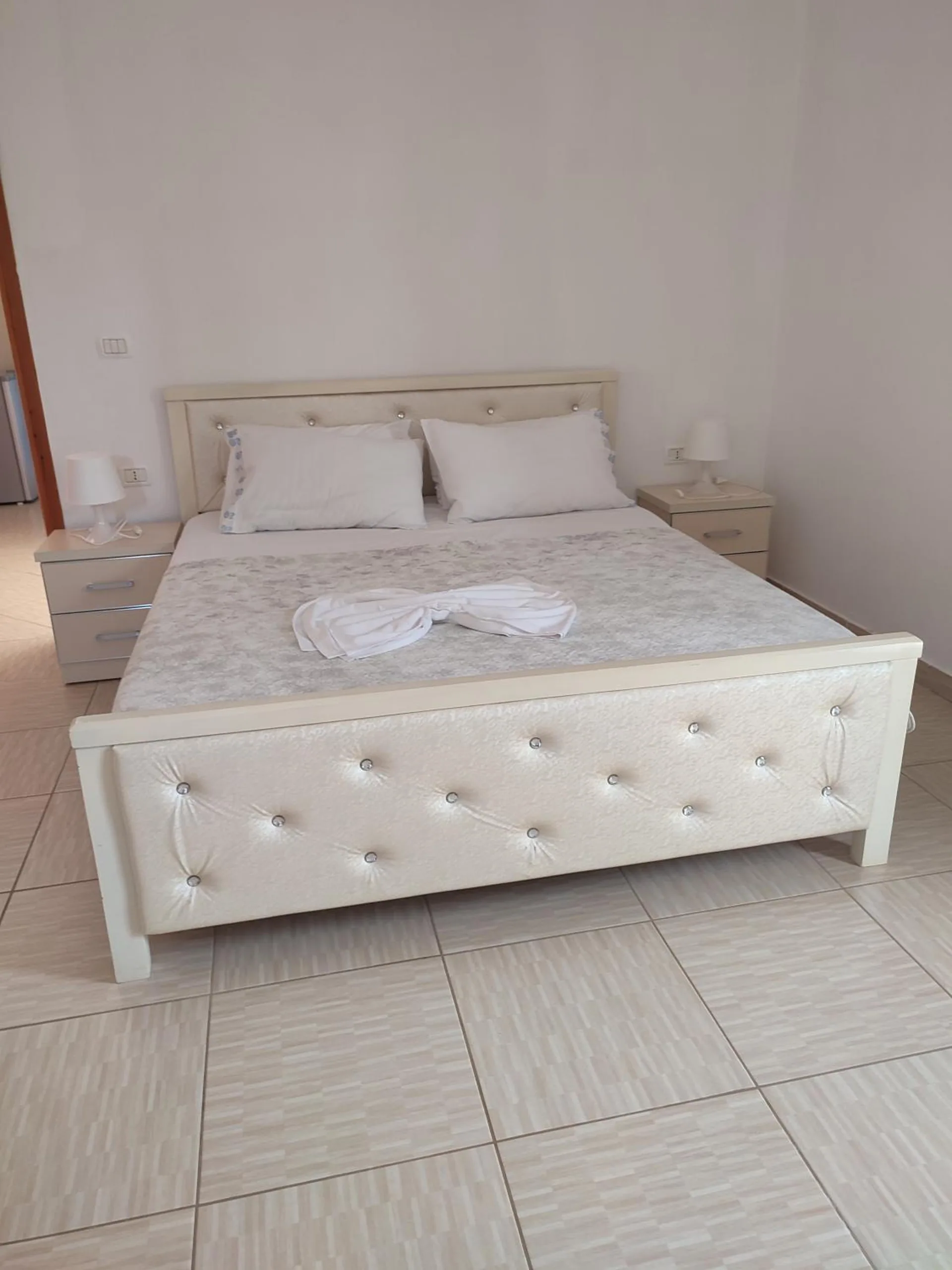 A white double bed with a buttoned headboard, towels folded on the cover, on a pale tiled floor