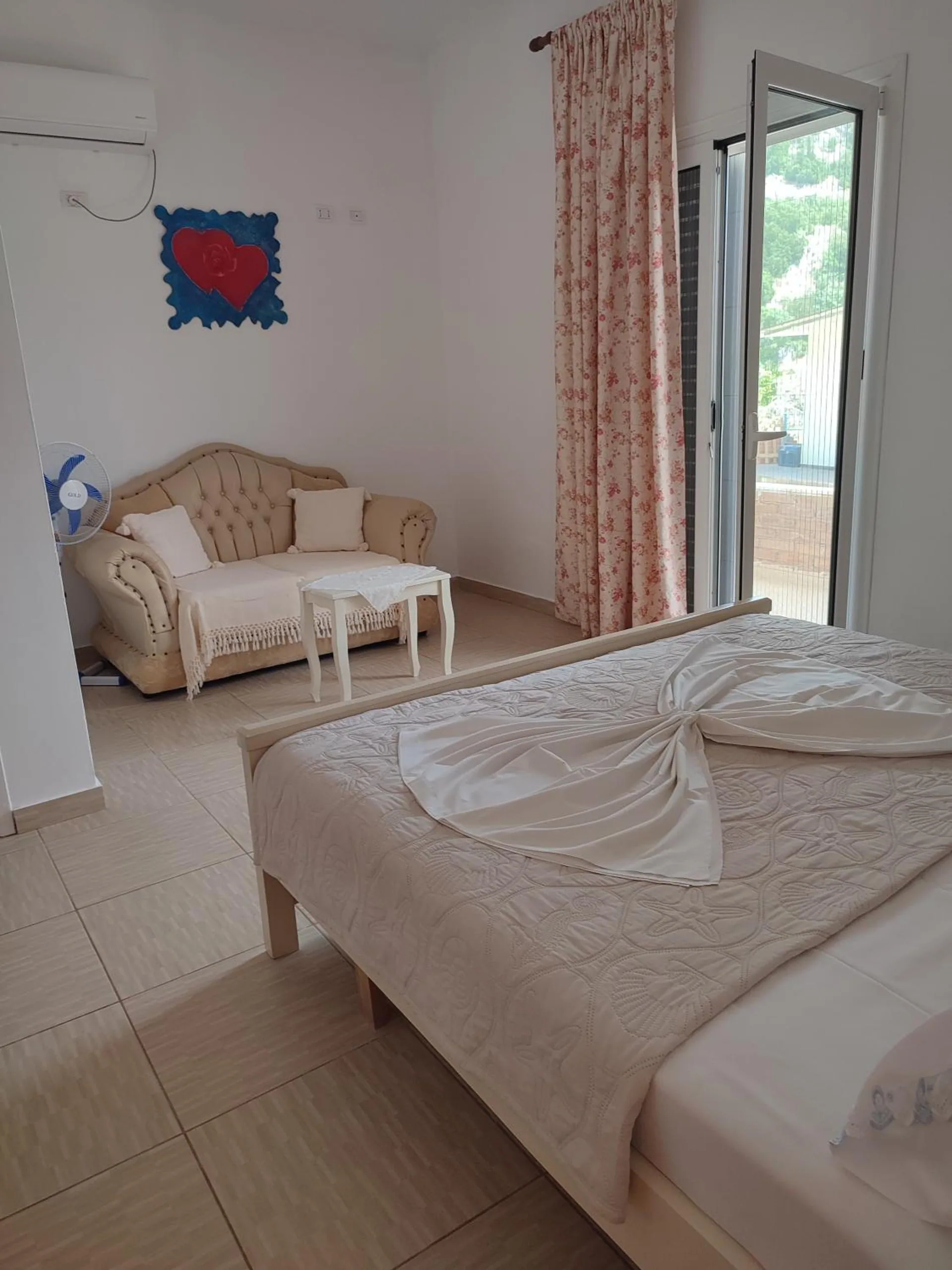 A double bed in a white room with a cream sofa, a small table and a curtained door open to the balcony