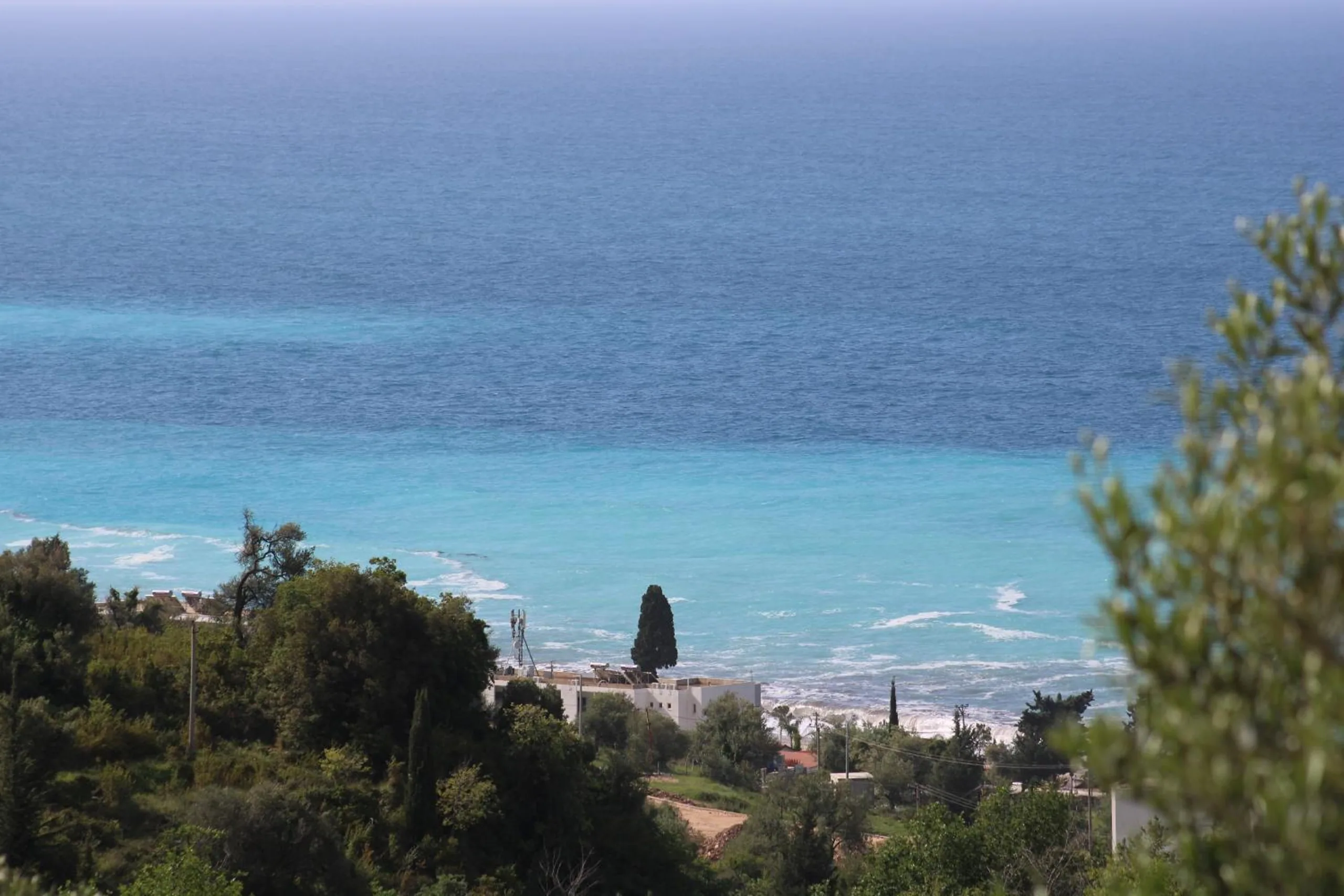 Turquoise sea and a long pale beach seen from the wooded hillside above, a single cypress among the trees