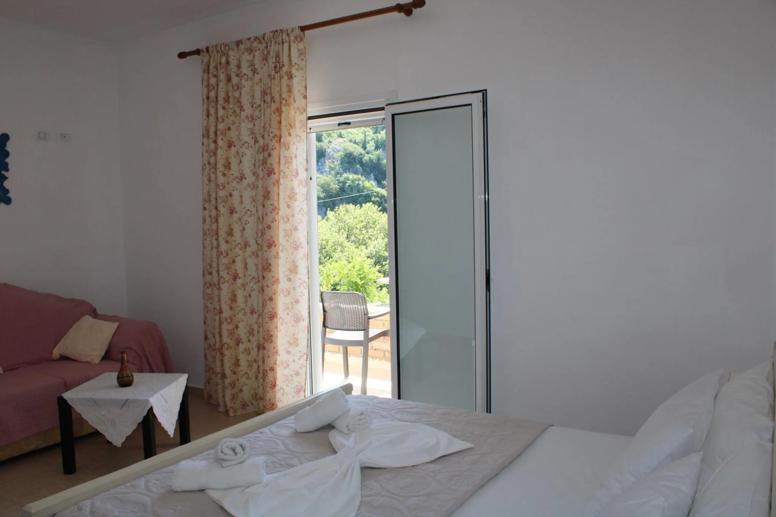 A double bed laid with towels, a pink sofa and a small table with a lace cloth, a door open to a balcony with chairs