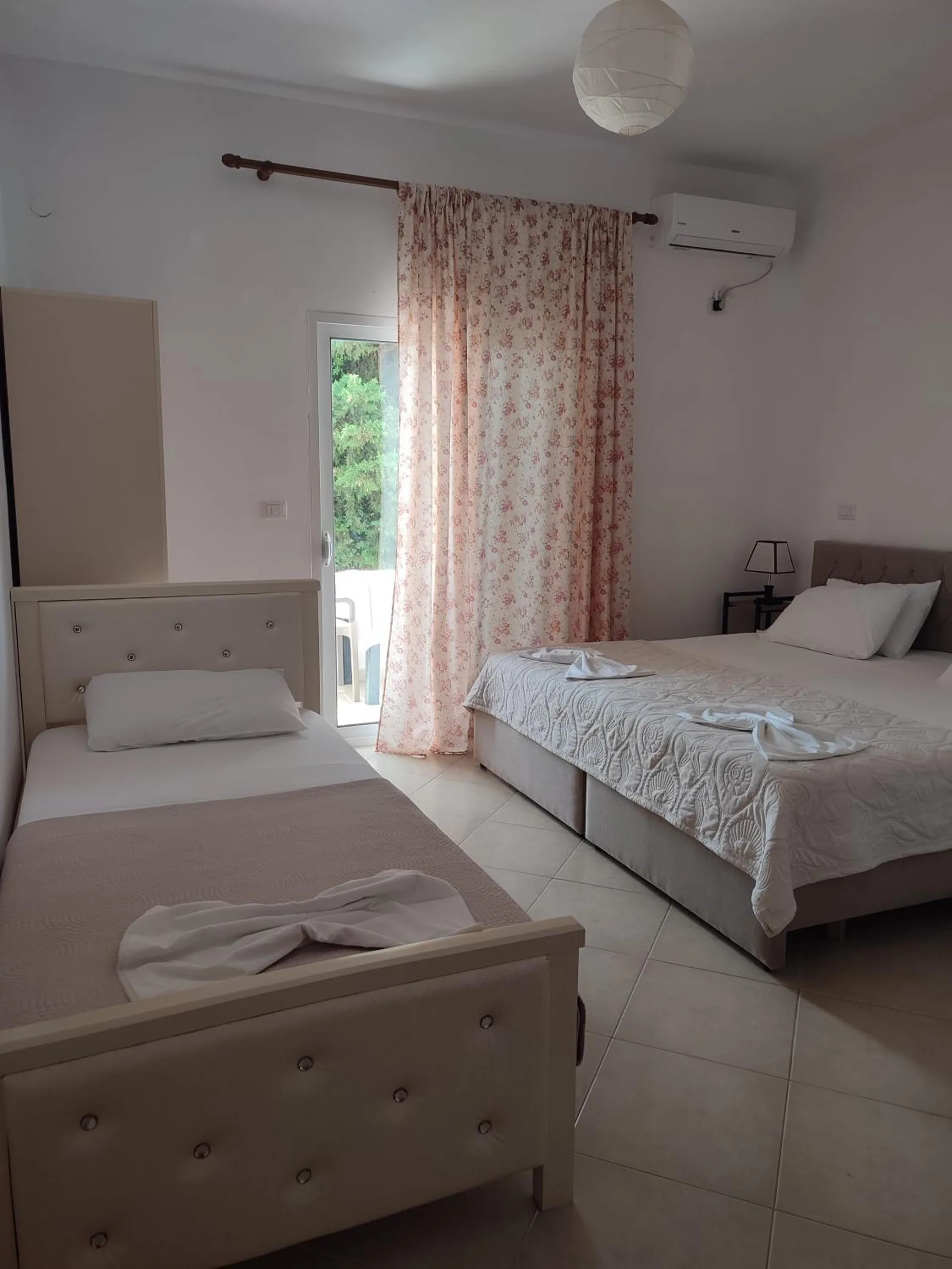 A single bed and a double bed side by side in a white room, an air-conditioning unit over the curtained balcony door