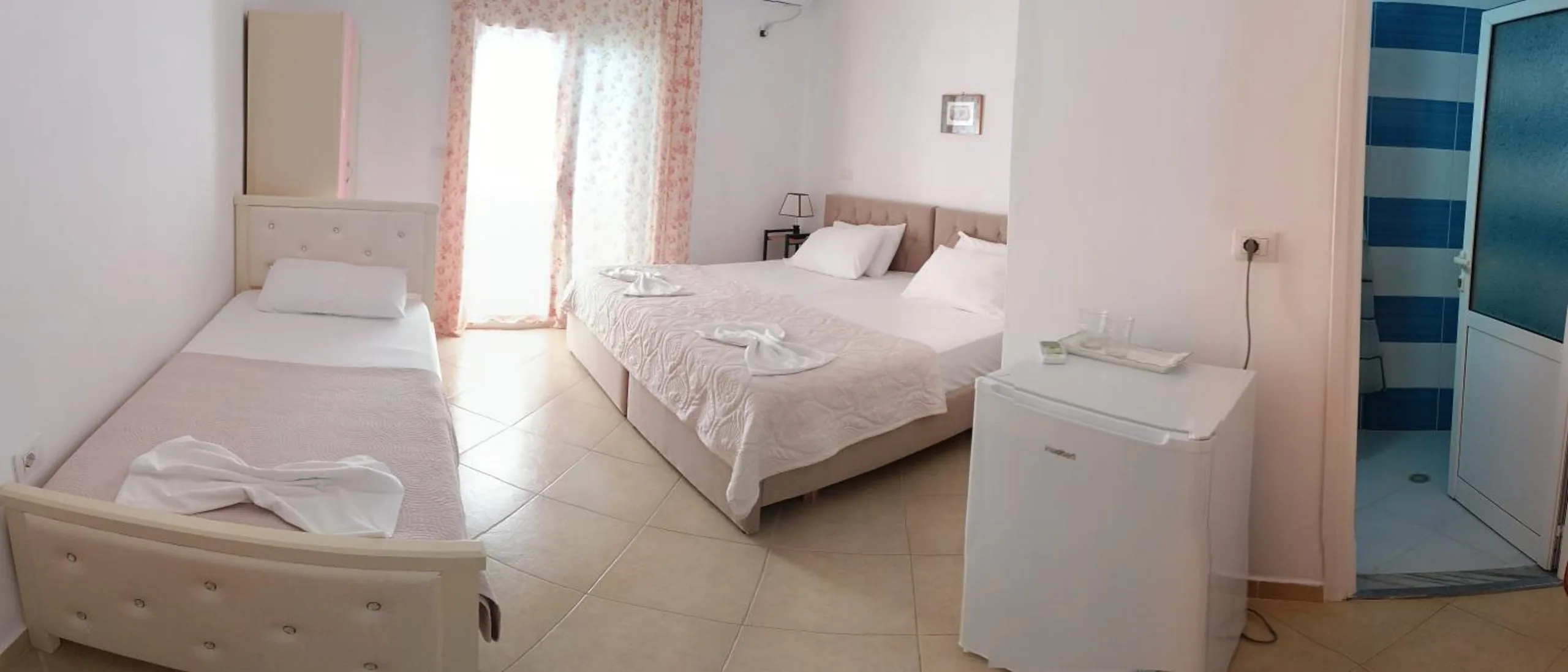 Panorama of a white room with a single bed, a double bed, a refrigerator and the door to a blue-tiled shower room