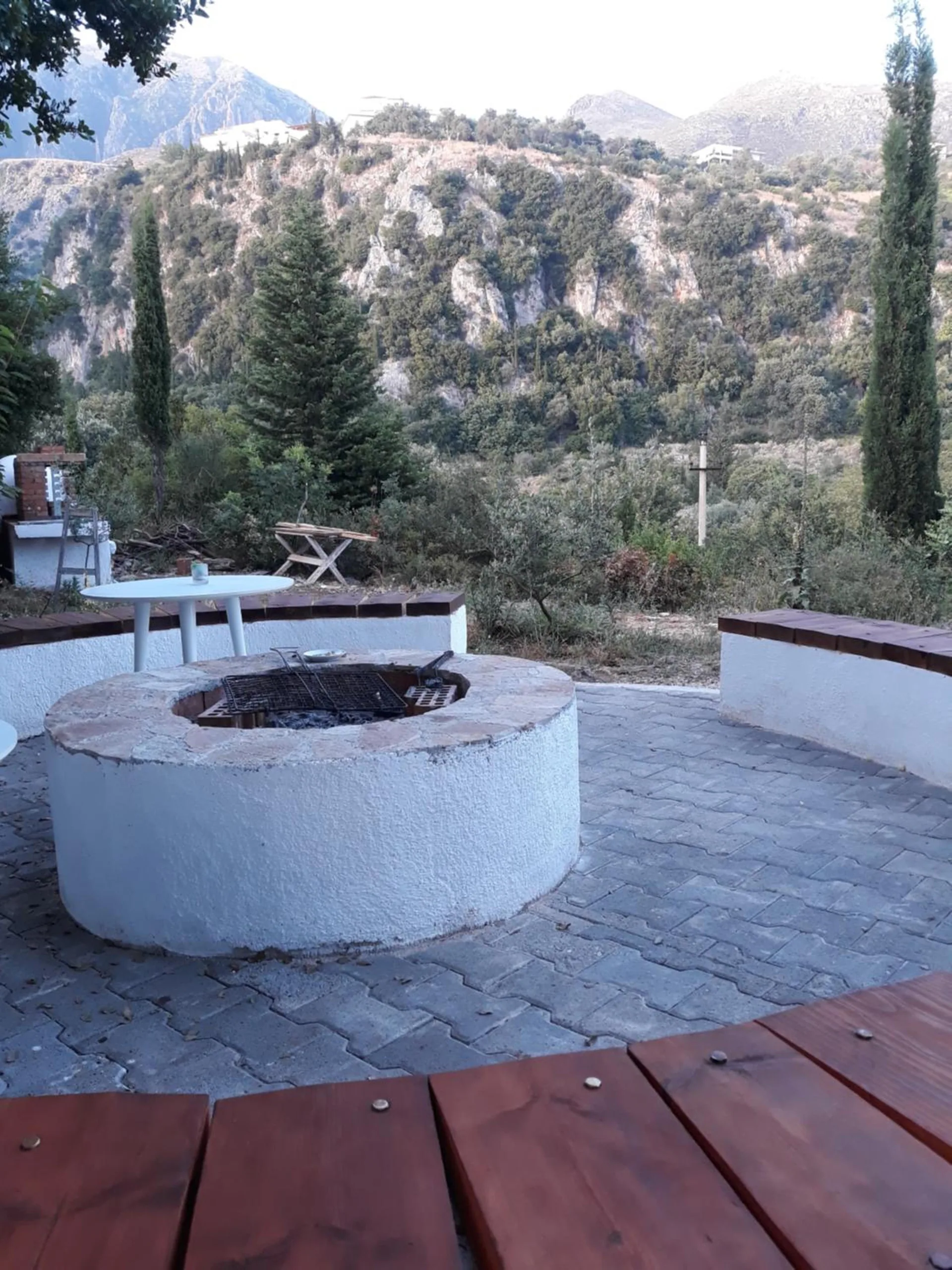 The whitewashed fire pit by day on a paved circle with timber bench seats, cypresses and a limestone hillside behind
