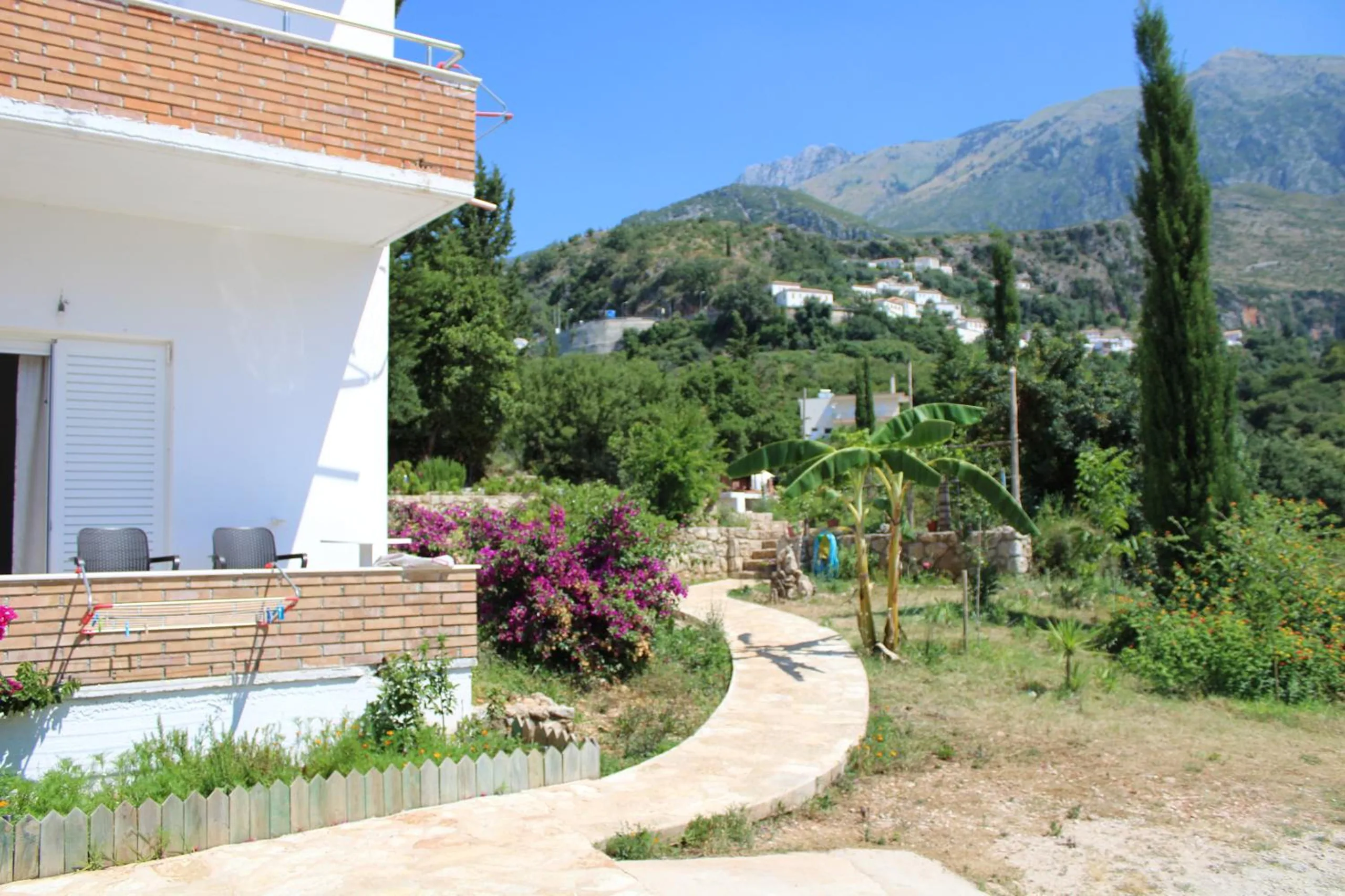 A curving paved path through the garden past the ground-floor terrace, a banana plant and a cypress, with white houses and mountains beyond