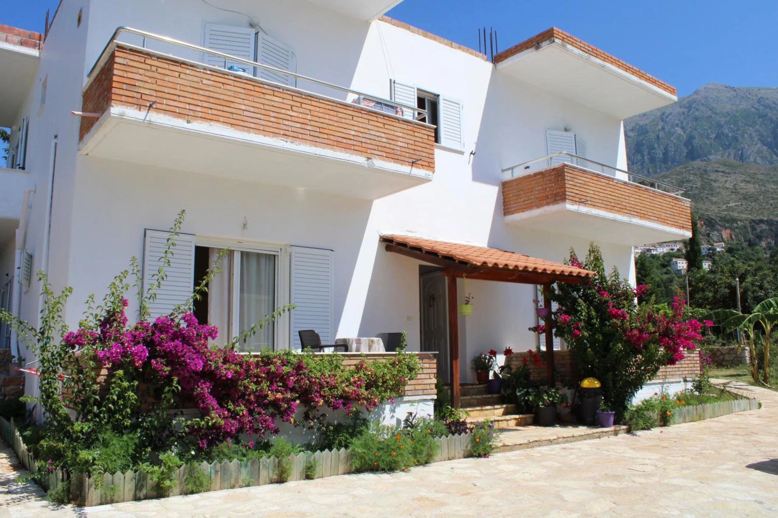 The white three-storey house with brick-faced balconies, a tiled pergola over the porch and magenta bougainvillea along the front, mountains behind