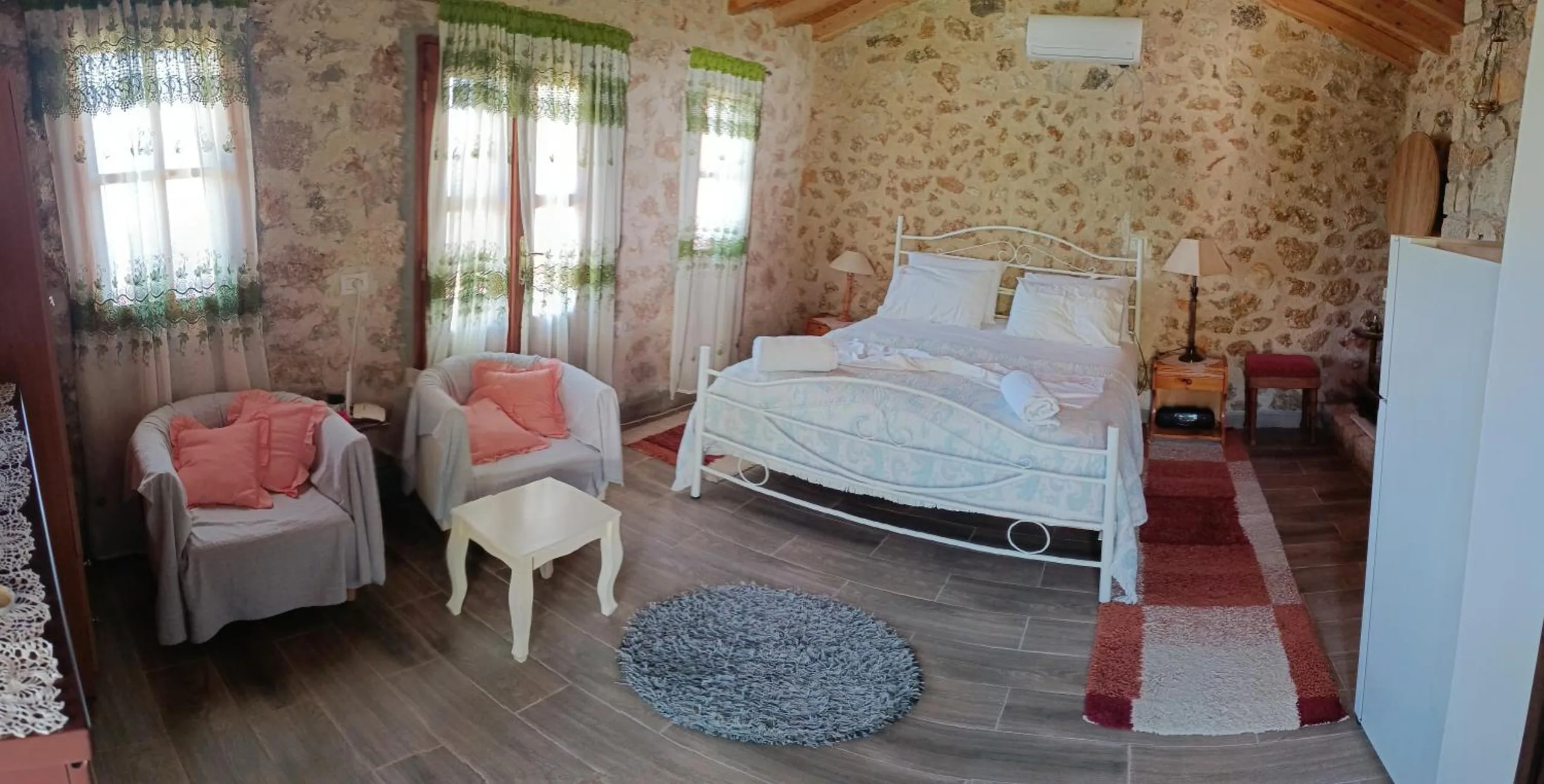 A stone-walled room with a white iron double bed, two armchairs with peach cushions, a red striped runner and curtained windows