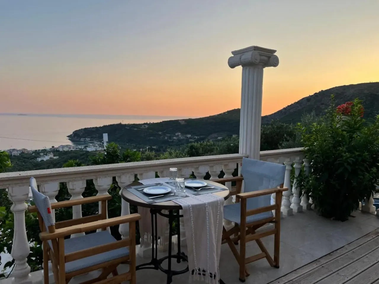 A laid table and two chairs on a terrace beside a fluted column with an Ionic capital, the bay and a pink sky beyond