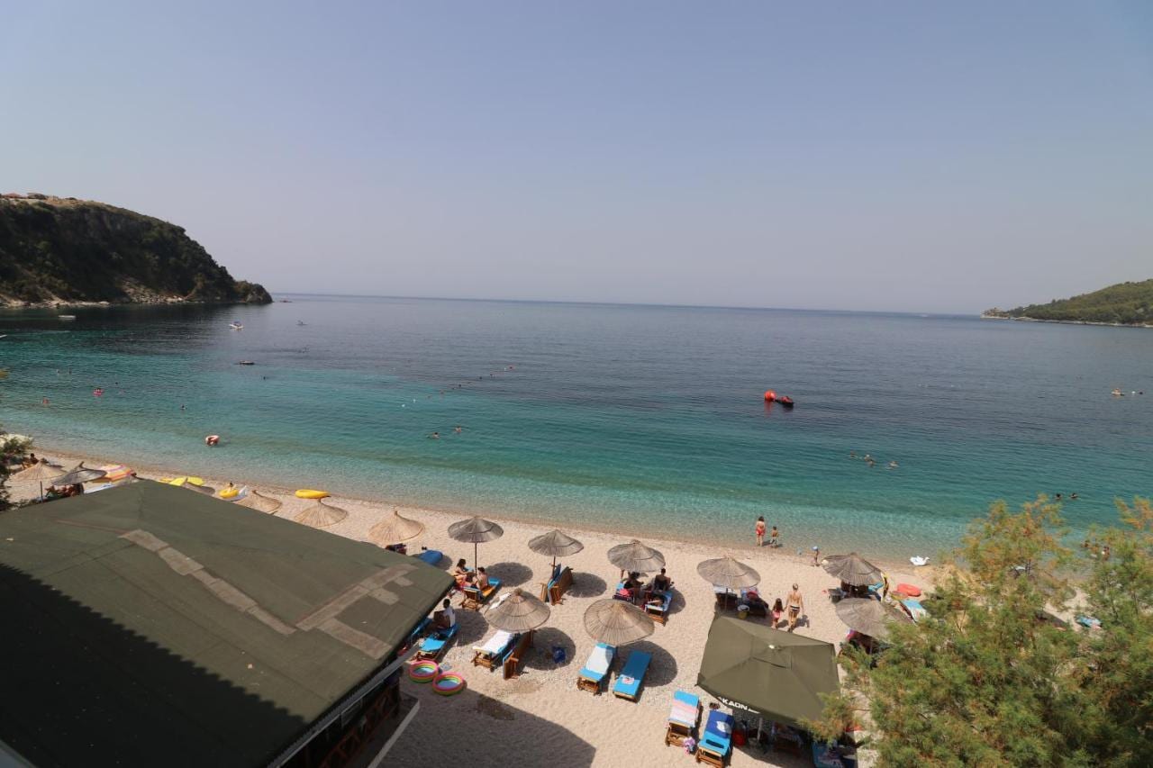The beach seen from an upper floor: umbrellas and sunbeds along the shingle, the clear bay and two wooded headlands