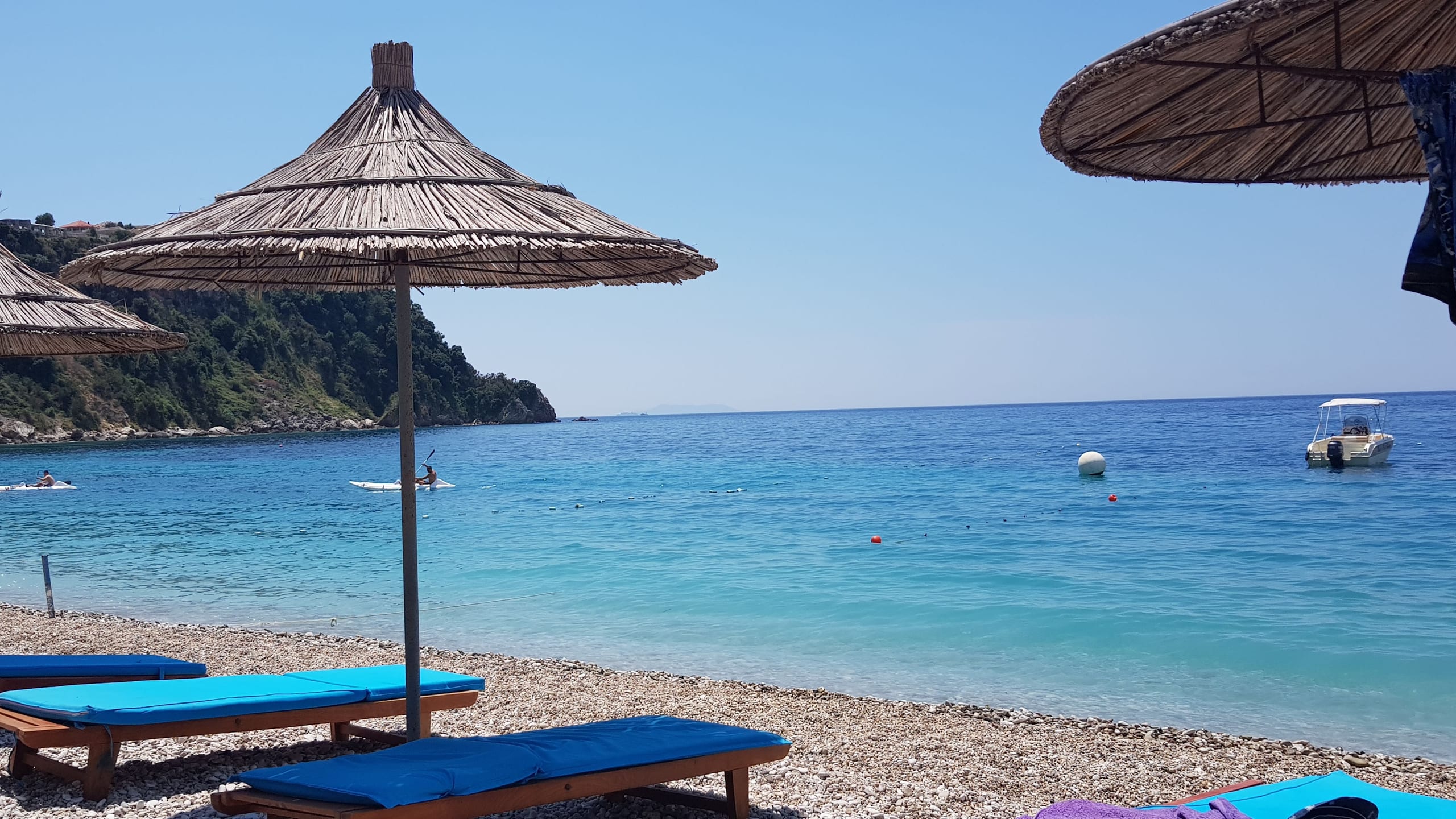 Thatched umbrellas and blue-cushioned sunbeds on the pebbles, with clear turquoise water, a moored motorboat and the wooded headland at the south end of the bay