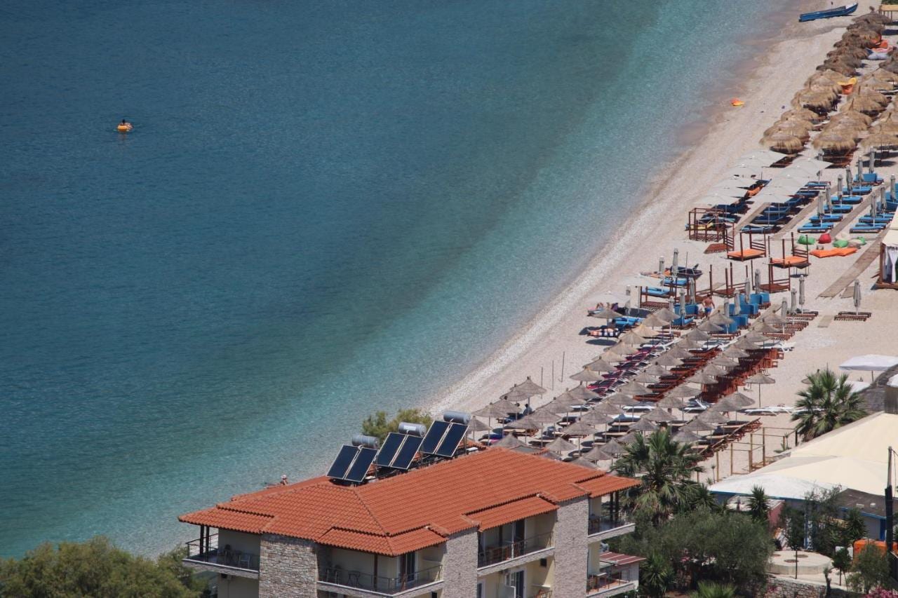 The hotel seen from the hillside: a terracotta-roofed building with stone piers and balconies at the edge of the shingle, a long line of umbrellas running north along the bay