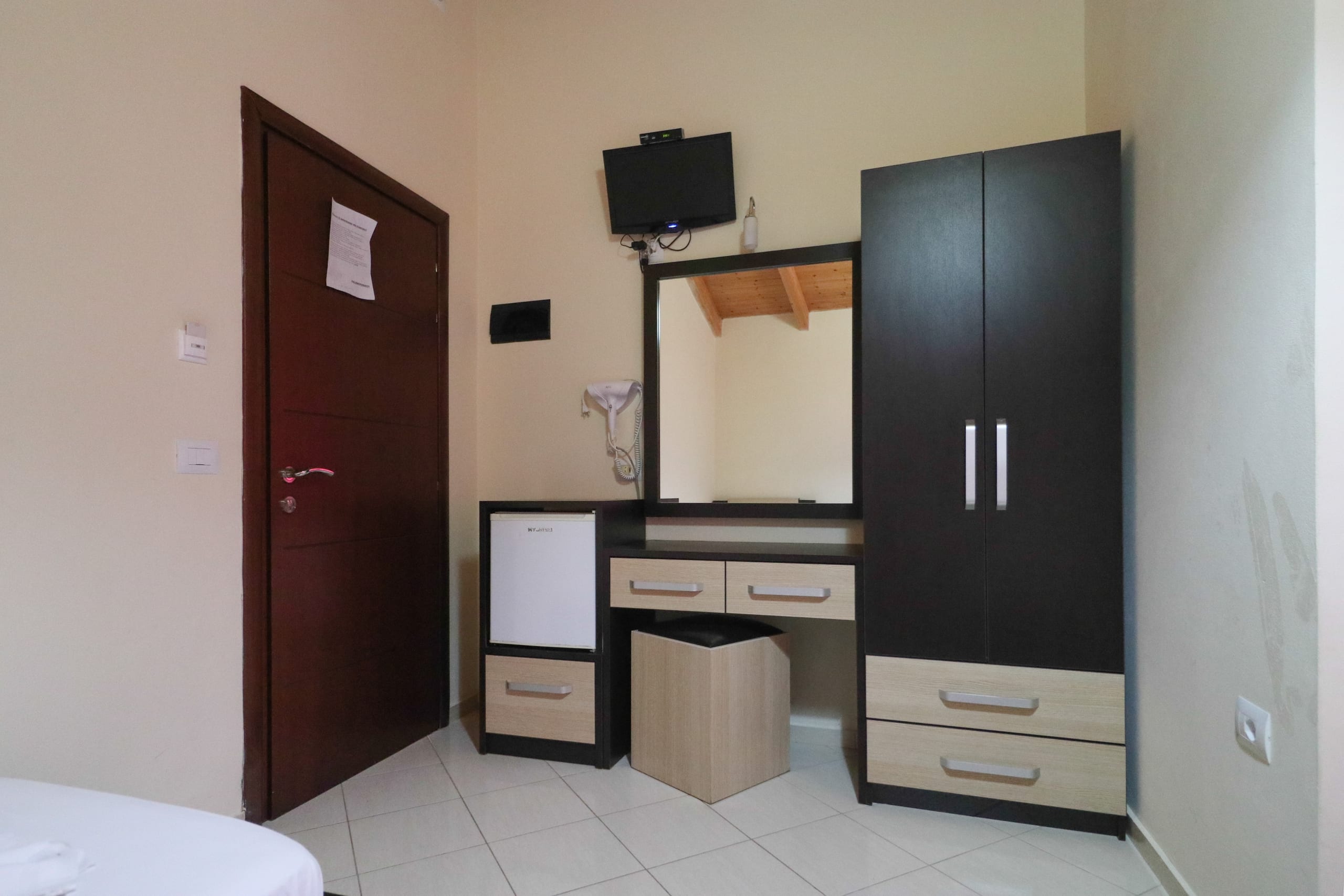 Inside a room's door: a dark wardrobe, a dressing table with mirror and hairdryer, a small fridge and a wall-mounted television