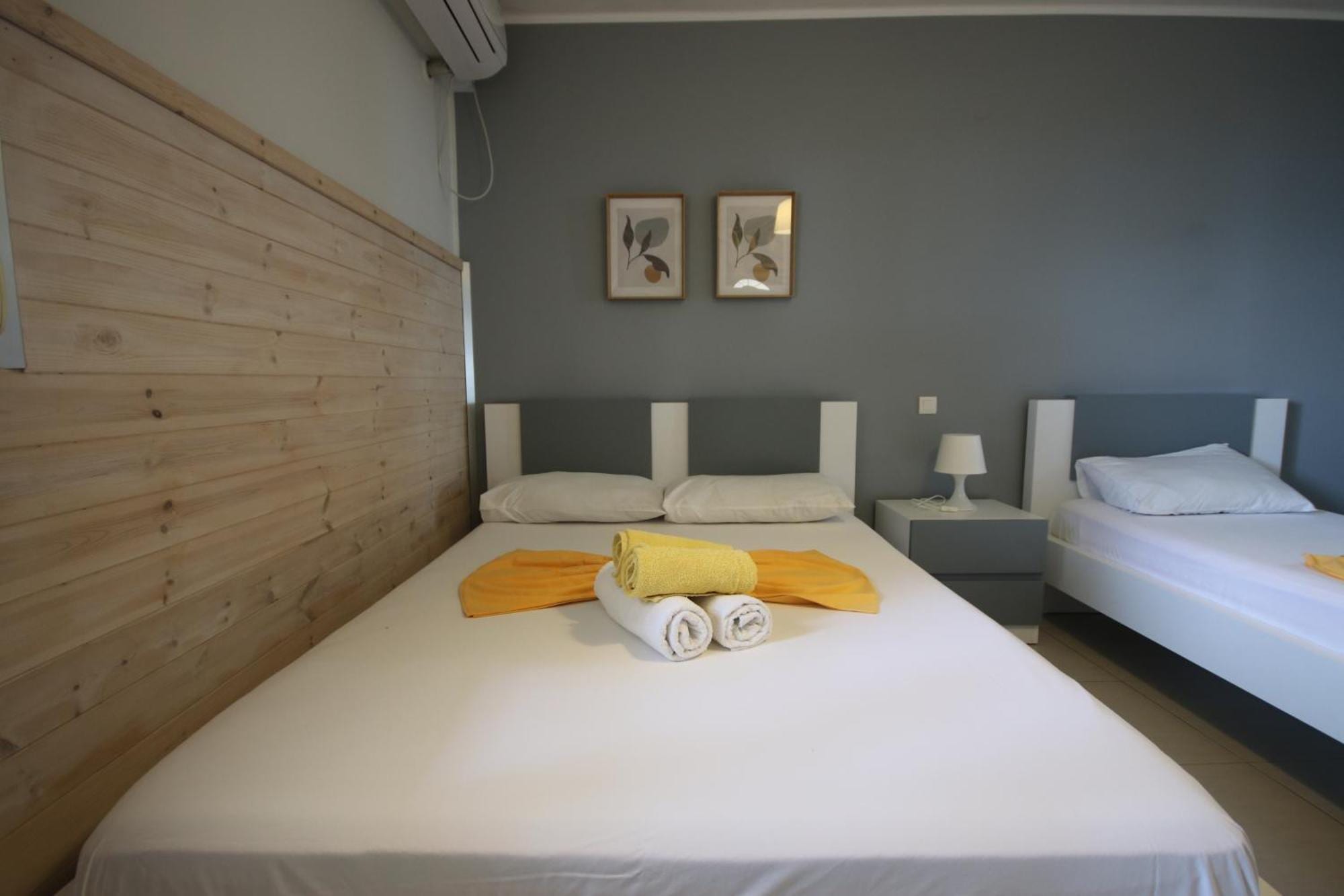 A grey-walled room with a double bed and a single, folded yellow towels, a pine-boarded wall and two small framed prints