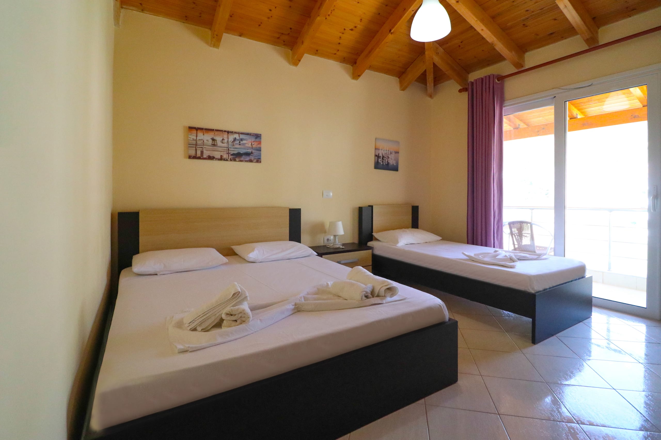 A room under a pitched timber ceiling with a double bed and a single bed, pale tiled floor and a balcony door with a purple curtain