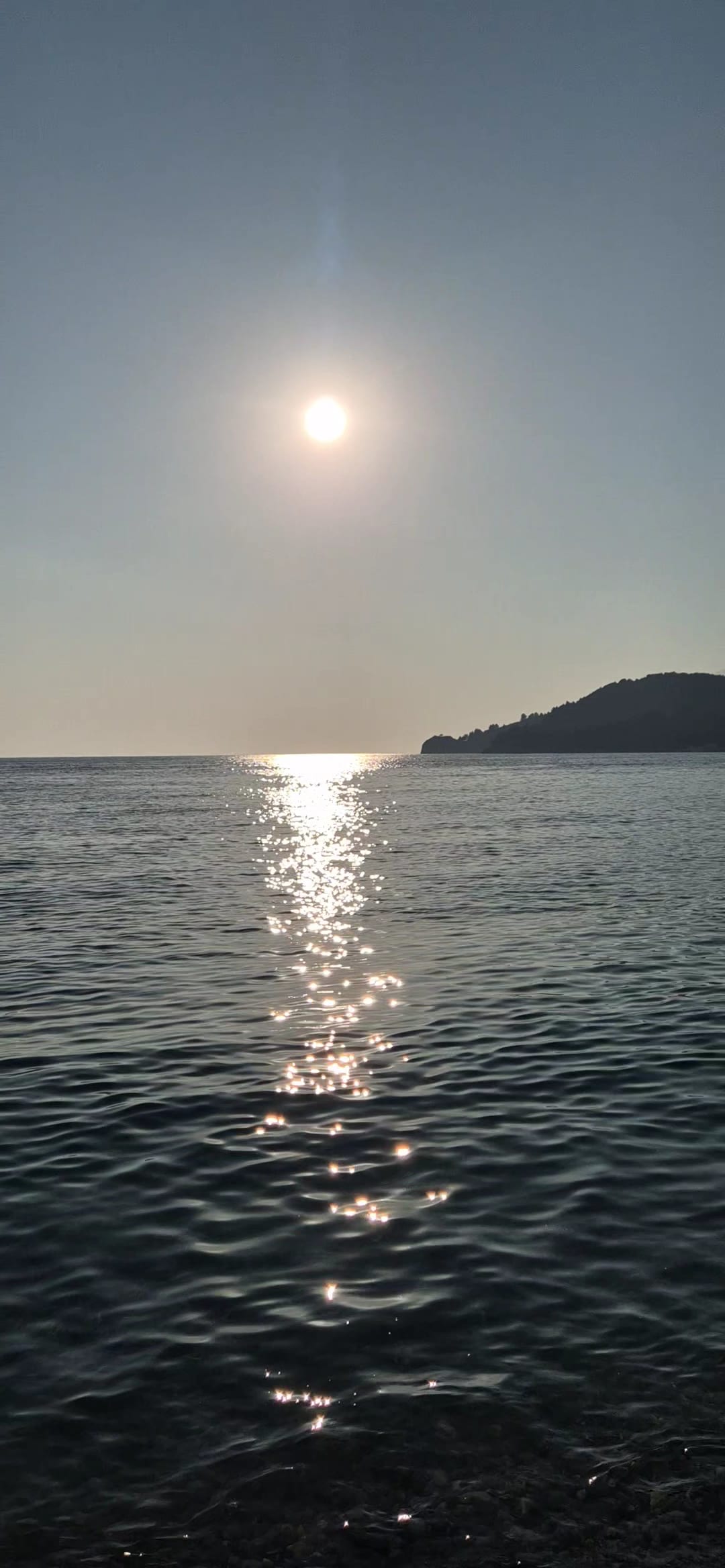 The sun low over a calm sea, its reflection running towards the shore, a dark headland on the right