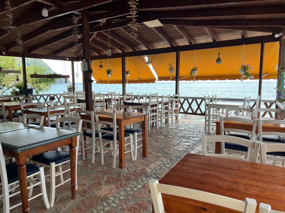 The restaurant terrace under a timber roof and a saffron awning, wooden tables and white chairs on a flagstone floor, the sea beyond the railing