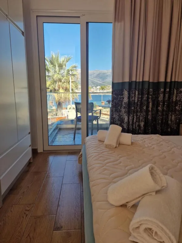A sliding balcony door in one of the rooms, a black mesh chair on the balcony, a palm, and the bay towards Himarë beyond