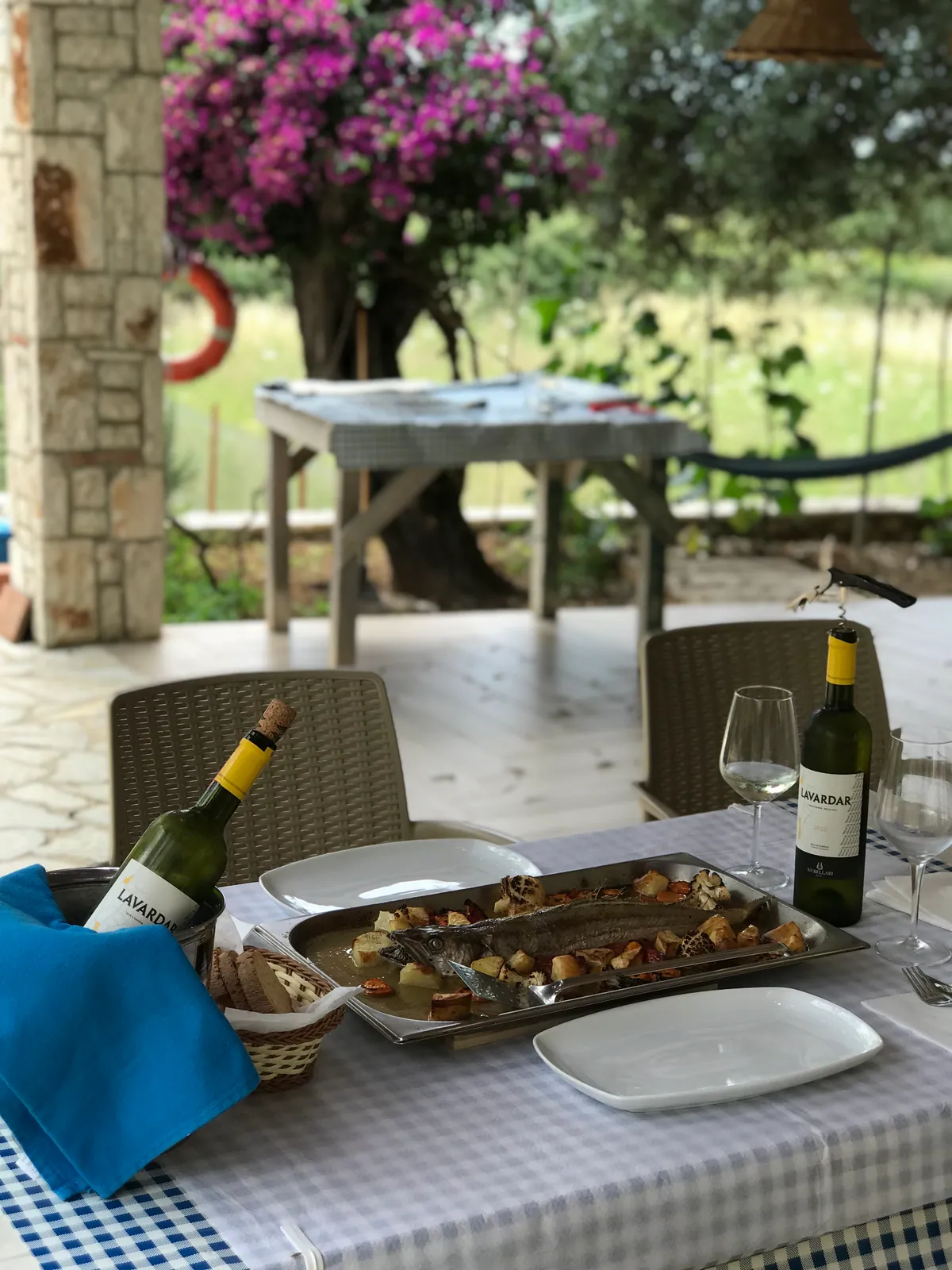 A whole fish baked in a tray with roast potatoes and vegetables on a gingham-covered table, two bottles of white wine, a stone pillar, a life ring and bougainvillea behind