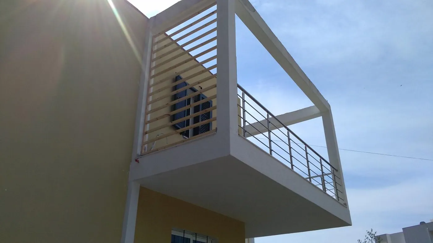 White concrete balcony frames with timber-slat screens and steel rails on a pale yellow wall, against a blue sky