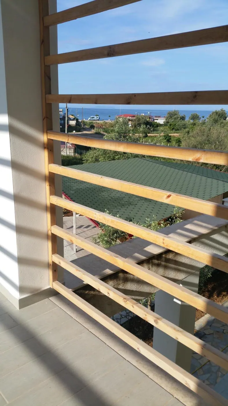 A balcony behind pale timber slats, looking over a green roof and olive trees to a strip of sea on the horizon