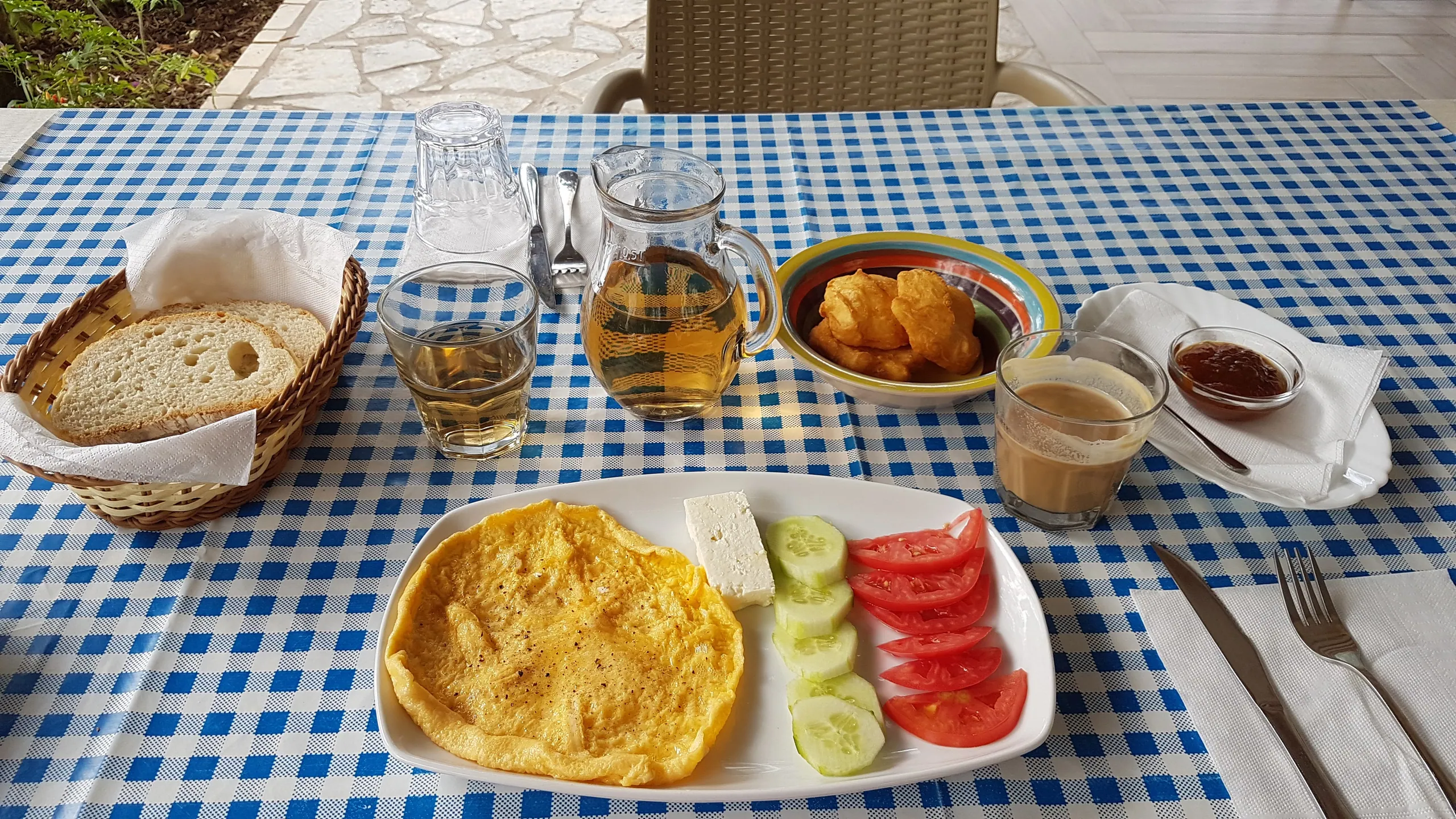 Breakfast on a blue and white gingham cloth: an omelette with feta, cucumber and tomato, a bowl of fried dough balls, bread in a basket, a jug of tea, coffee and a dish of jam