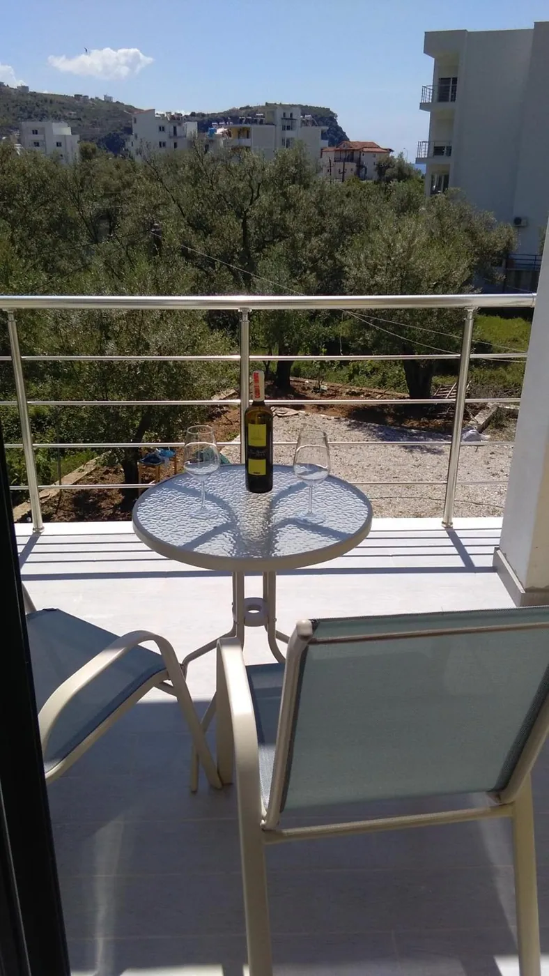 A balcony with two chairs and a round glass table with a bottle of wine, the steel rail looking over olive trees to the town on the hill