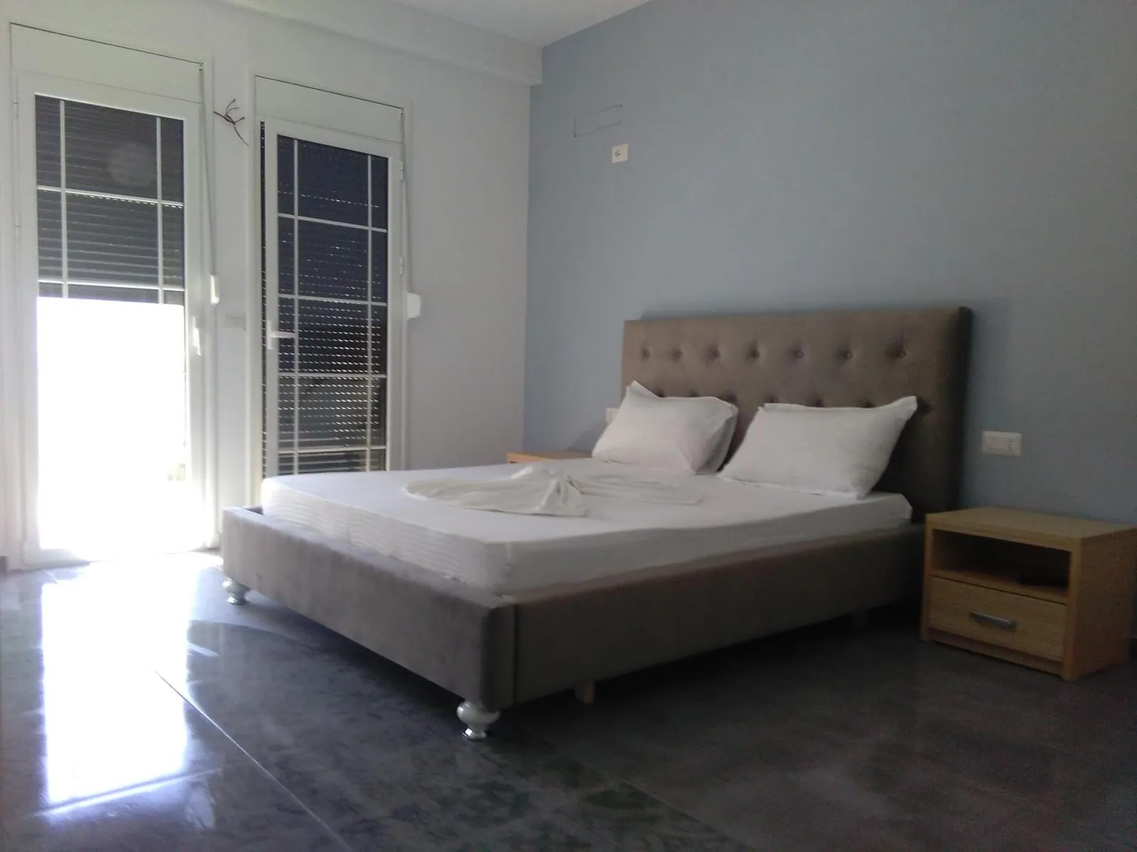A double bed with a grey buttoned headboard and white linen in a room with grey patterned floor tiles, a pale wood bedside table and glazed doors with shutters