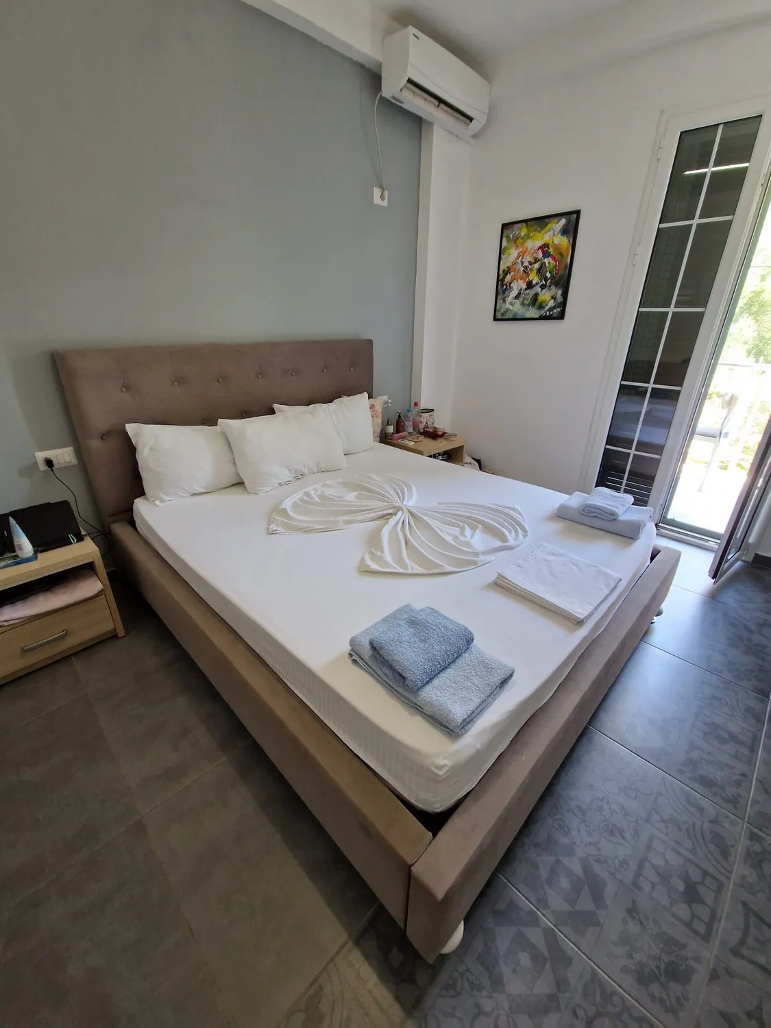 A double bed with an upholstered headboard and white linen folded into a bow, blue towels at the foot, a bedside table, a painting on the white wall and a glazed balcony door