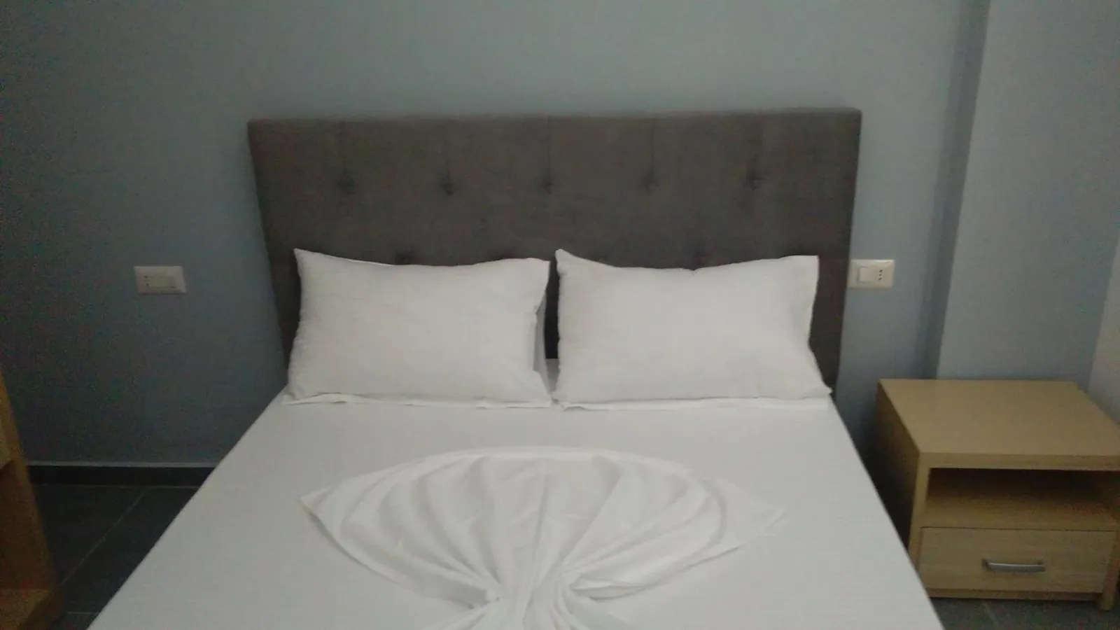 A double bed with a grey upholstered headboard and white linen against a pale blue wall, a light wood bedside table beside it