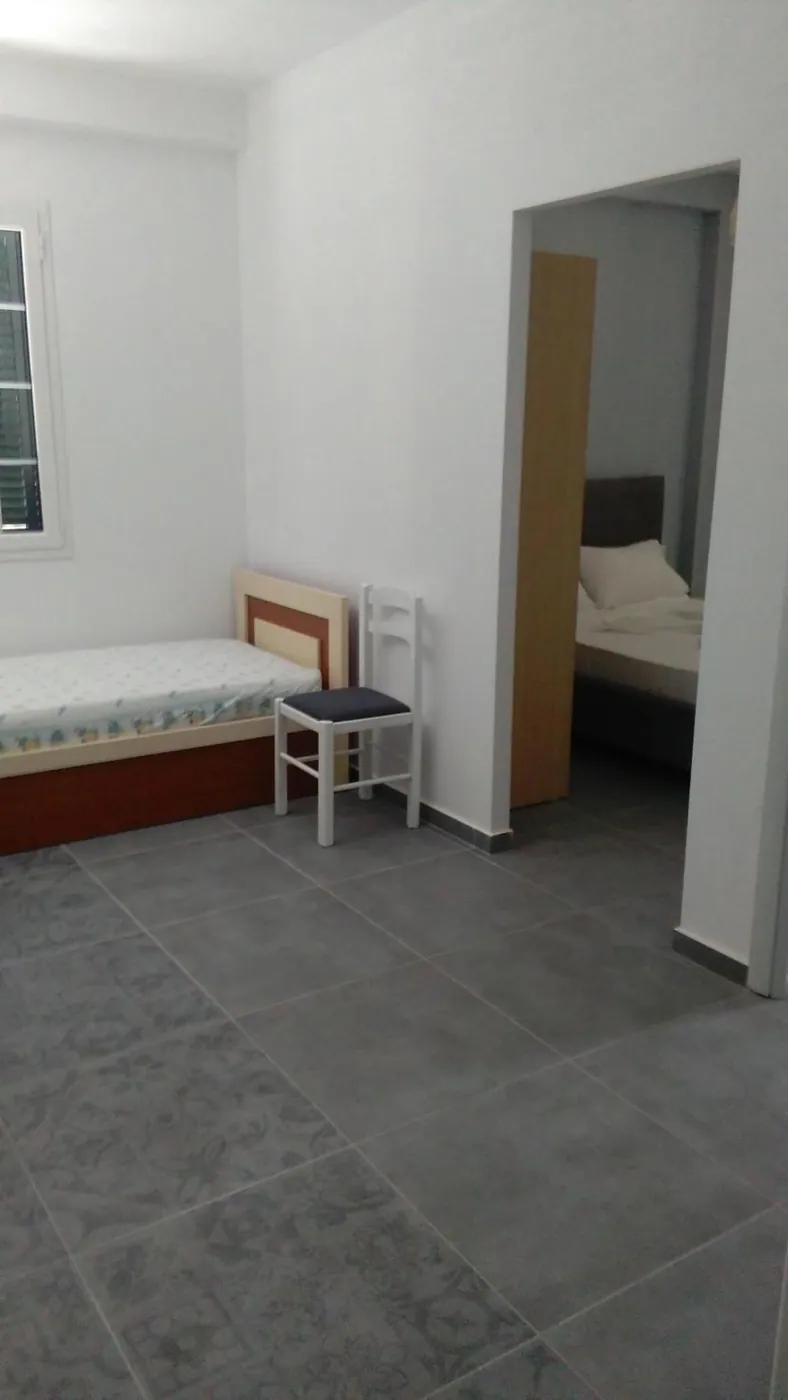 A single bed with a red frame and a chair against a white wall, grey floor tiles, seen through a doorway