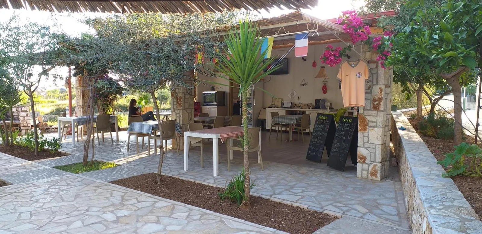 The open garden kitchen and bar under a reed roof, gingham-covered tables on the flagstones, two chalk menu boards, flags and a bougainvillea on a stone pillar