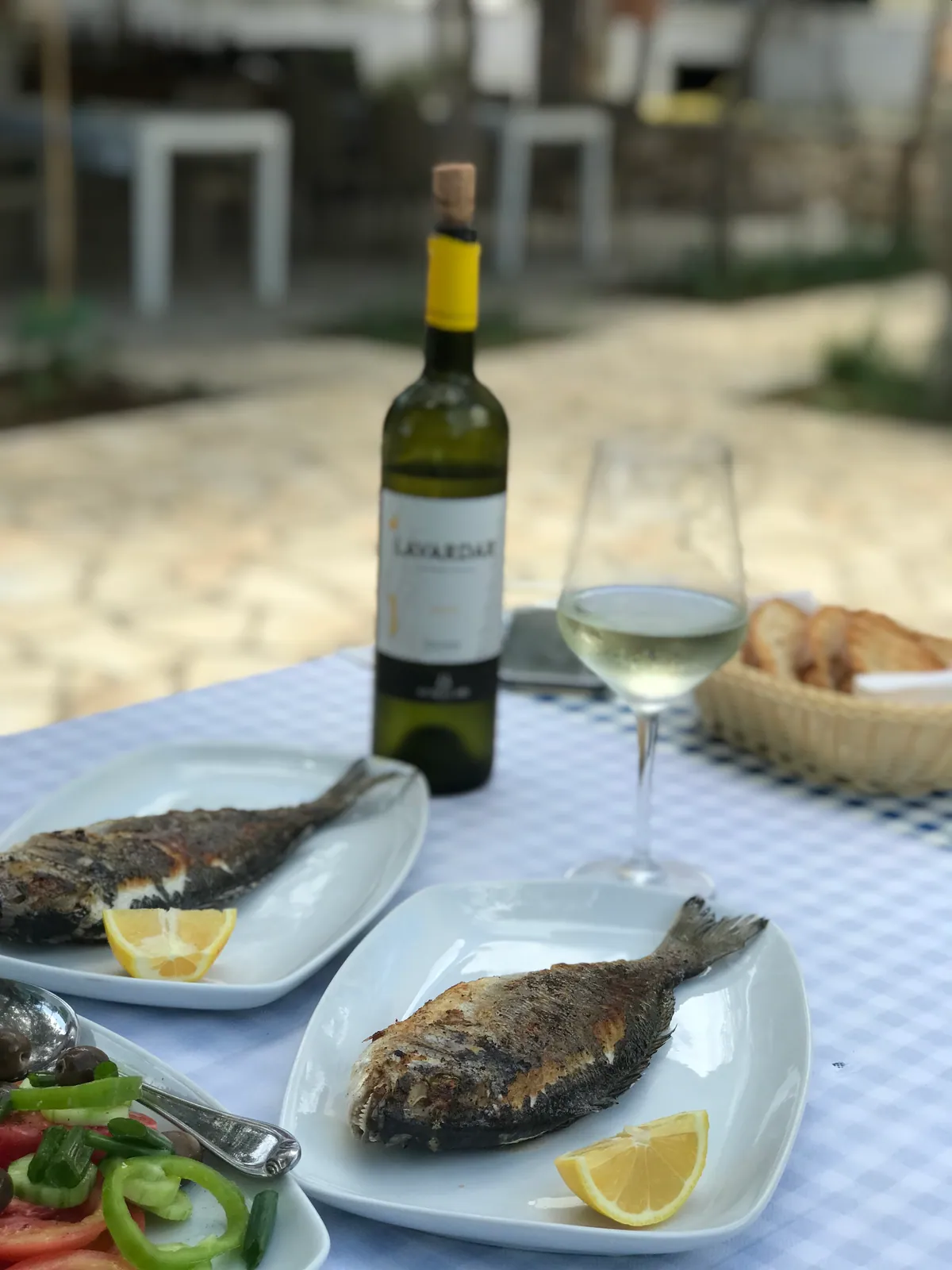 Two grilled sea bream with lemon on white plates, a salad, a basket of toasted bread and a glass and bottle of white wine on a blue gingham cloth, the flagstone terrace behind