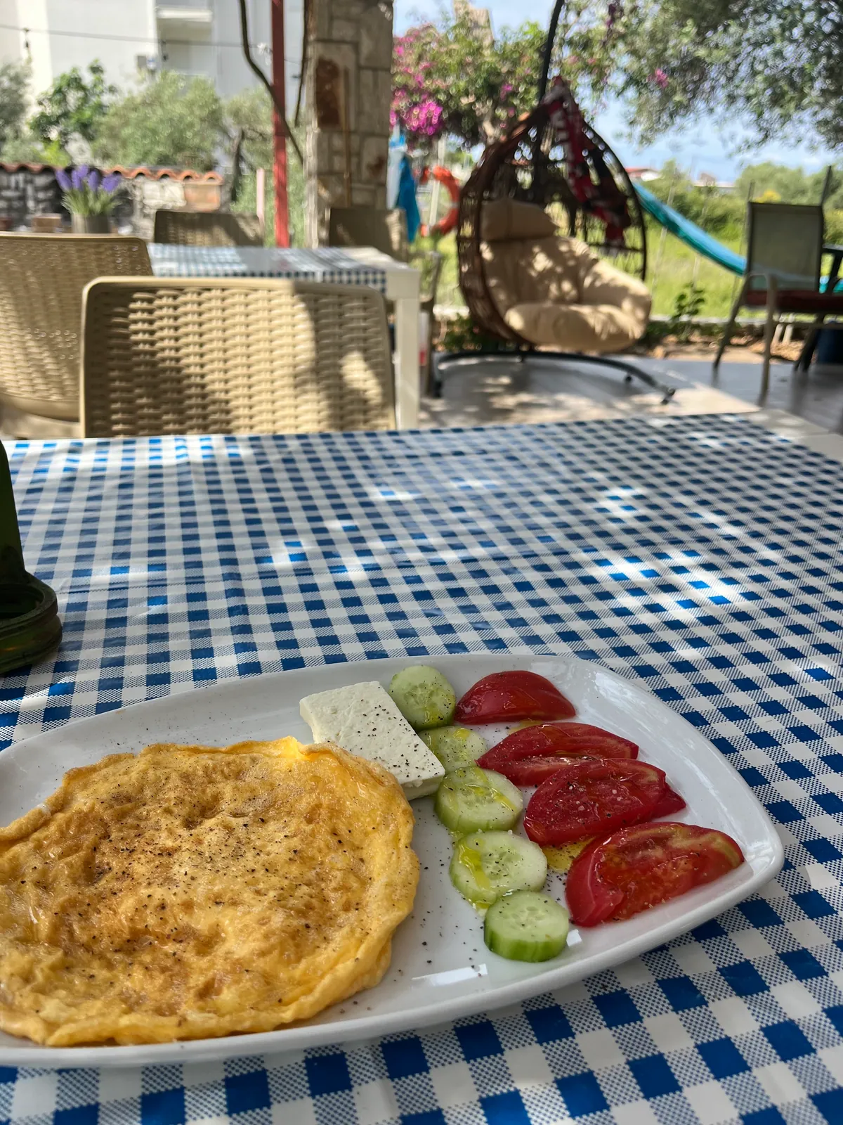An omelette with feta, sliced tomato and cucumber on a white plate on a blue and white gingham tablecloth, a hammock and the garden behind