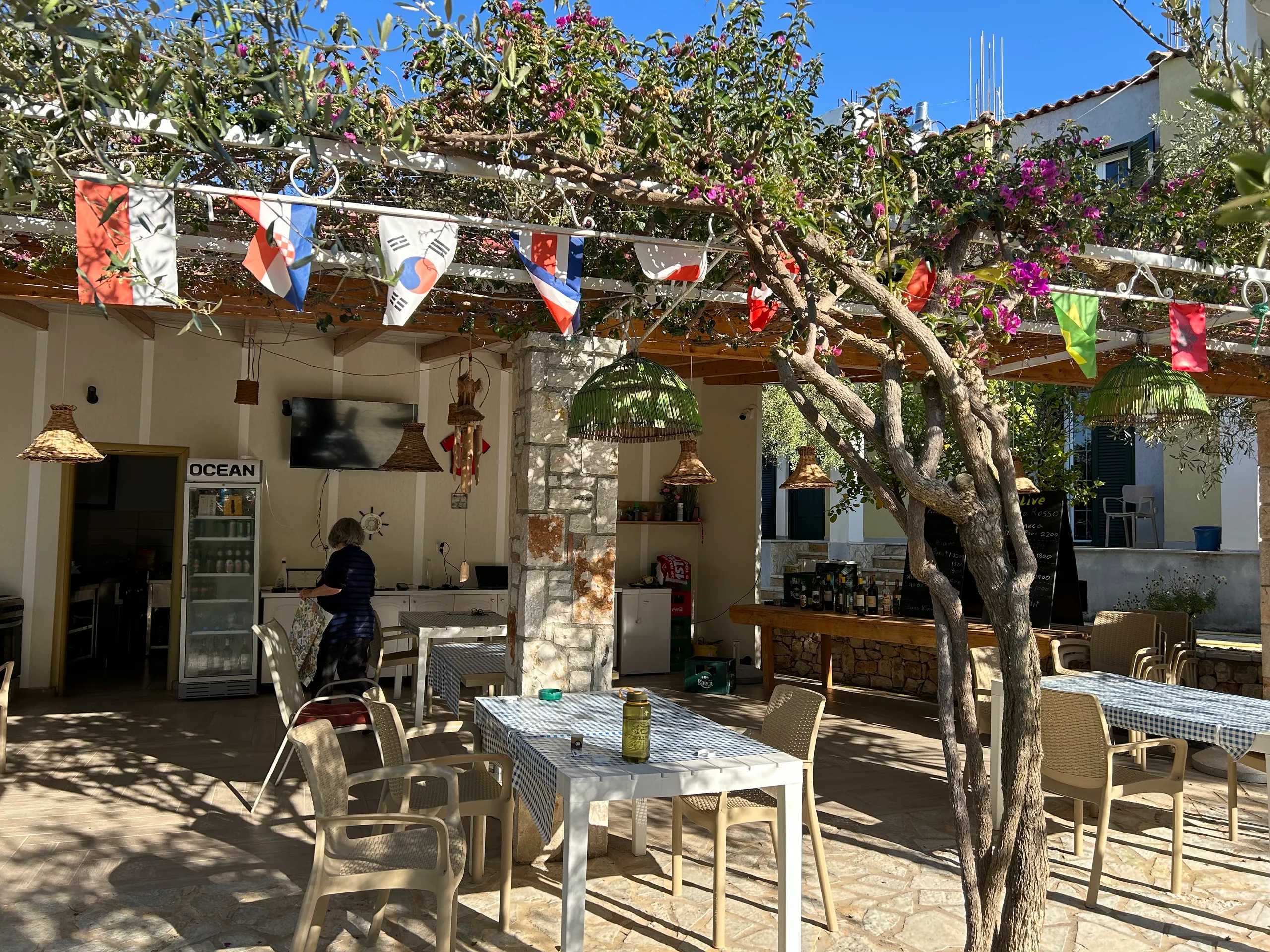 A bougainvillea-covered pergola hung with flags of many countries and woven lampshades over gingham-covered tables, a stone pillar, the bar counter and a drinks fridge behind