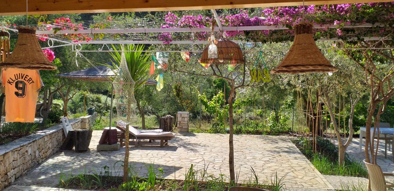 Wicker lampshades hanging under a pergola covered in pink and magenta bougainvillea, a sun lounger on the flagstone terrace and the vegetable garden beyond