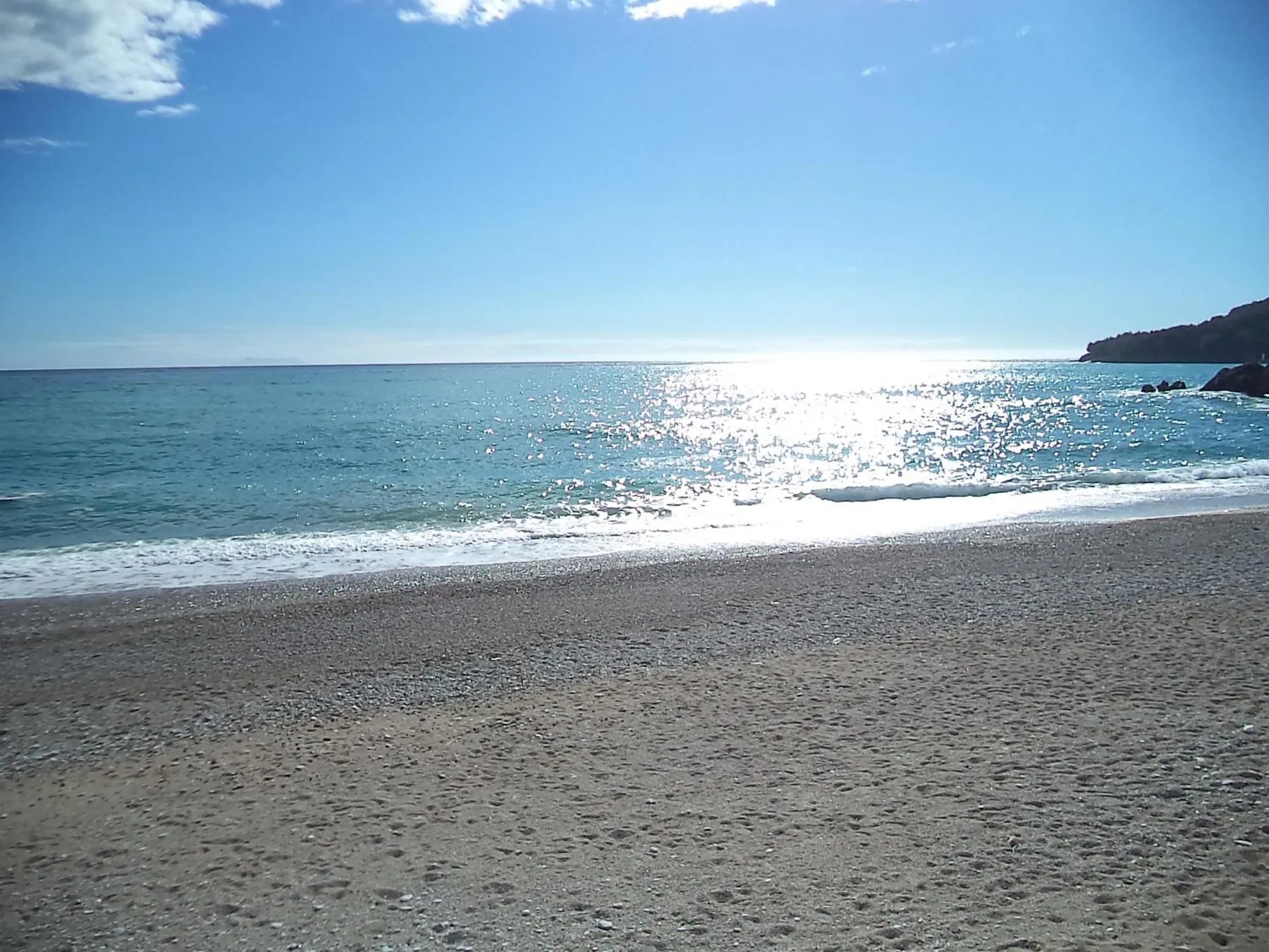 A shingle beach and a low breaking wave under a sun glinting on the sea, a rocky headland on the right