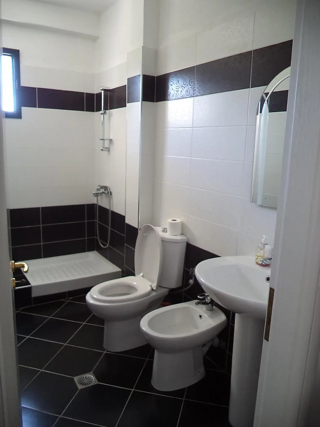 A shower room with a shower tray, basin, toilet and bidet, white wall tiles with a dark band and dark floor tiles
