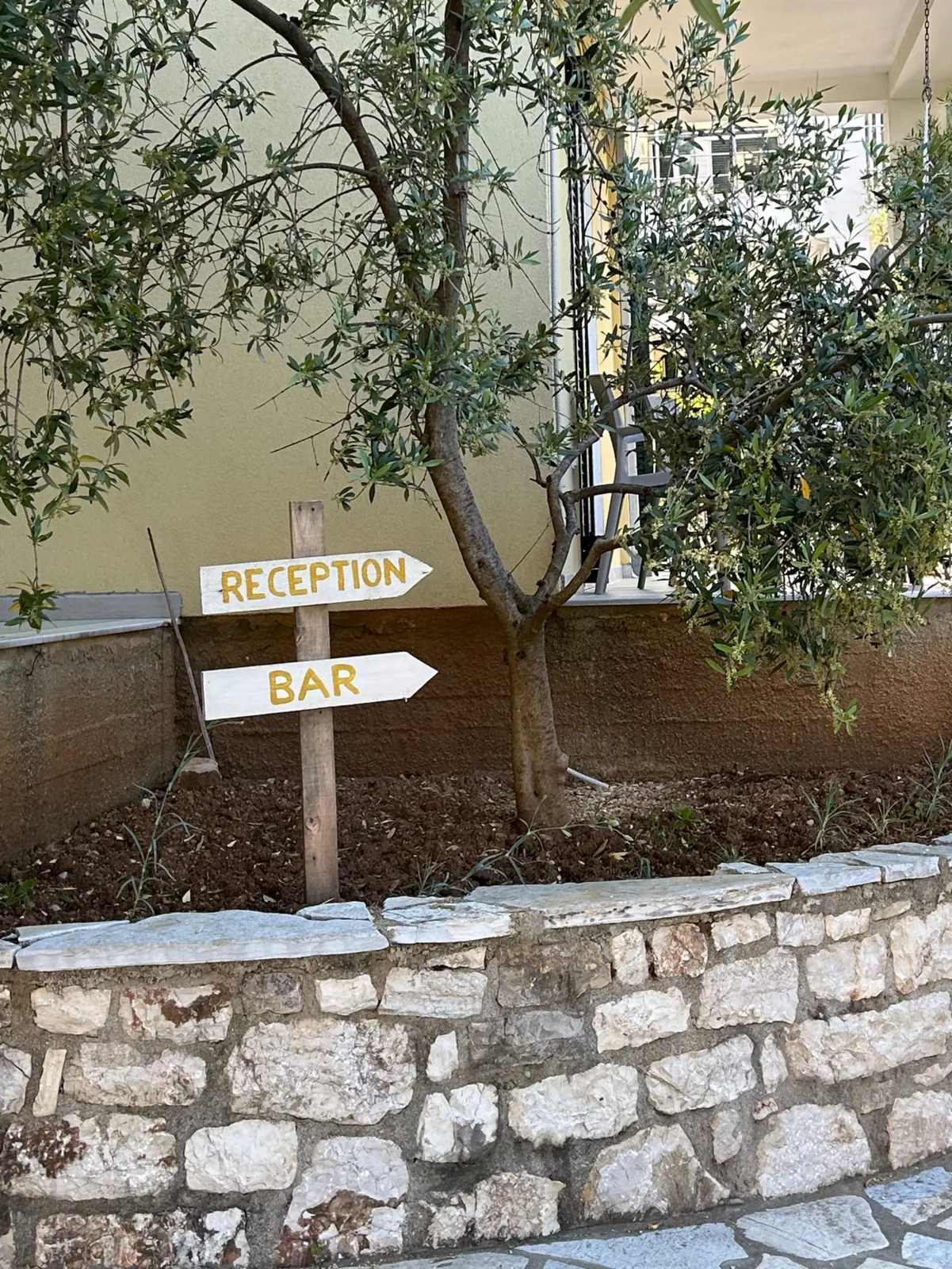 A young olive tree in a curved dry-stone planter in front of a pale yellow house, a hand-painted wooden signpost pointing to Reception and Bar
