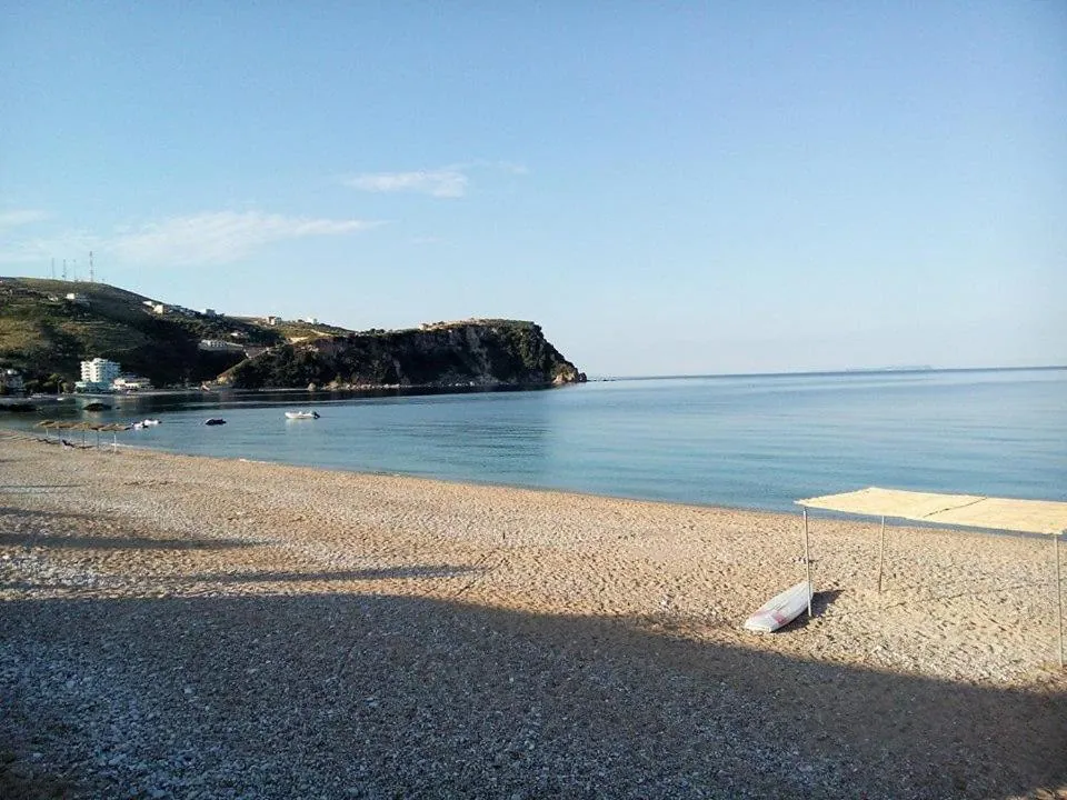 A curve of shingle beach in early light, a shade sail and a small boat on the stones, a green headland with a harbour at its foot across calm water