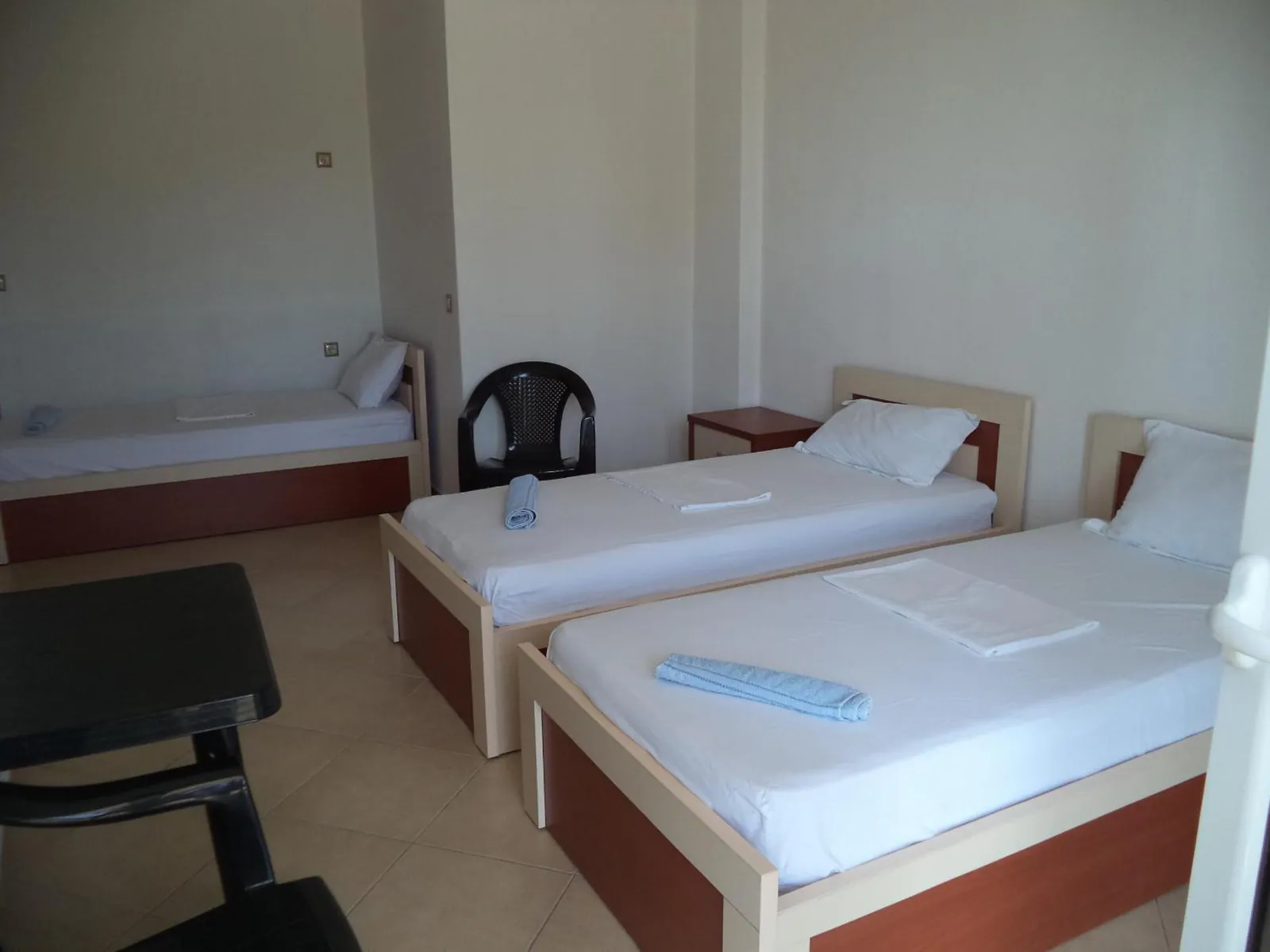 Three single beds with white linen and pale wood frames in a white room, blue towels on the pillows and a dark bedside table
