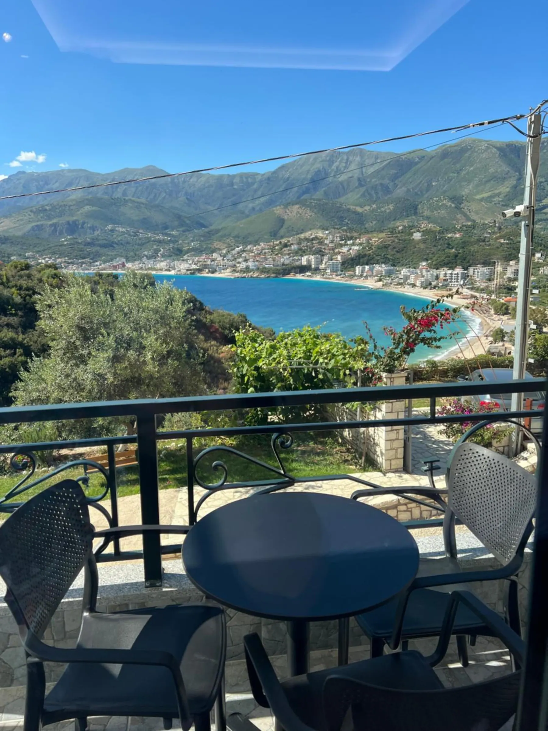 An upstairs balcony with a dark table and chairs, and beyond the rail the bay, the town and a bougainvillea in the garden below
