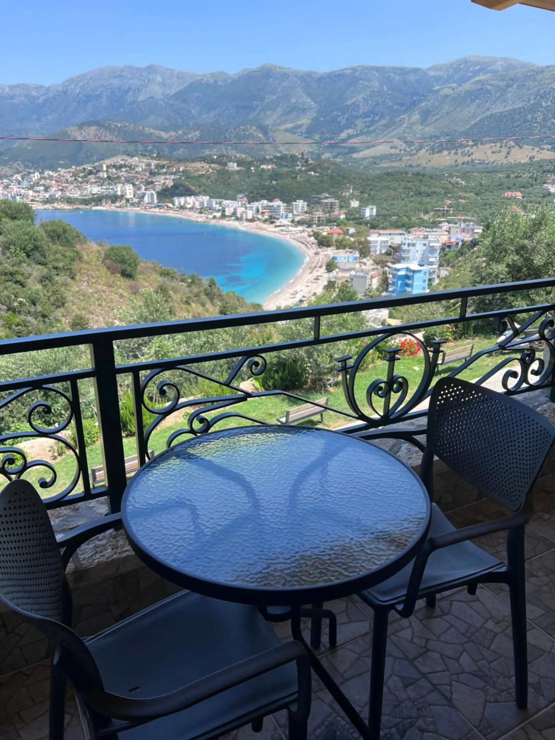 A glass table and two chairs on an upstairs balcony, and below the iron rail the whole curve of Potam bay and the town