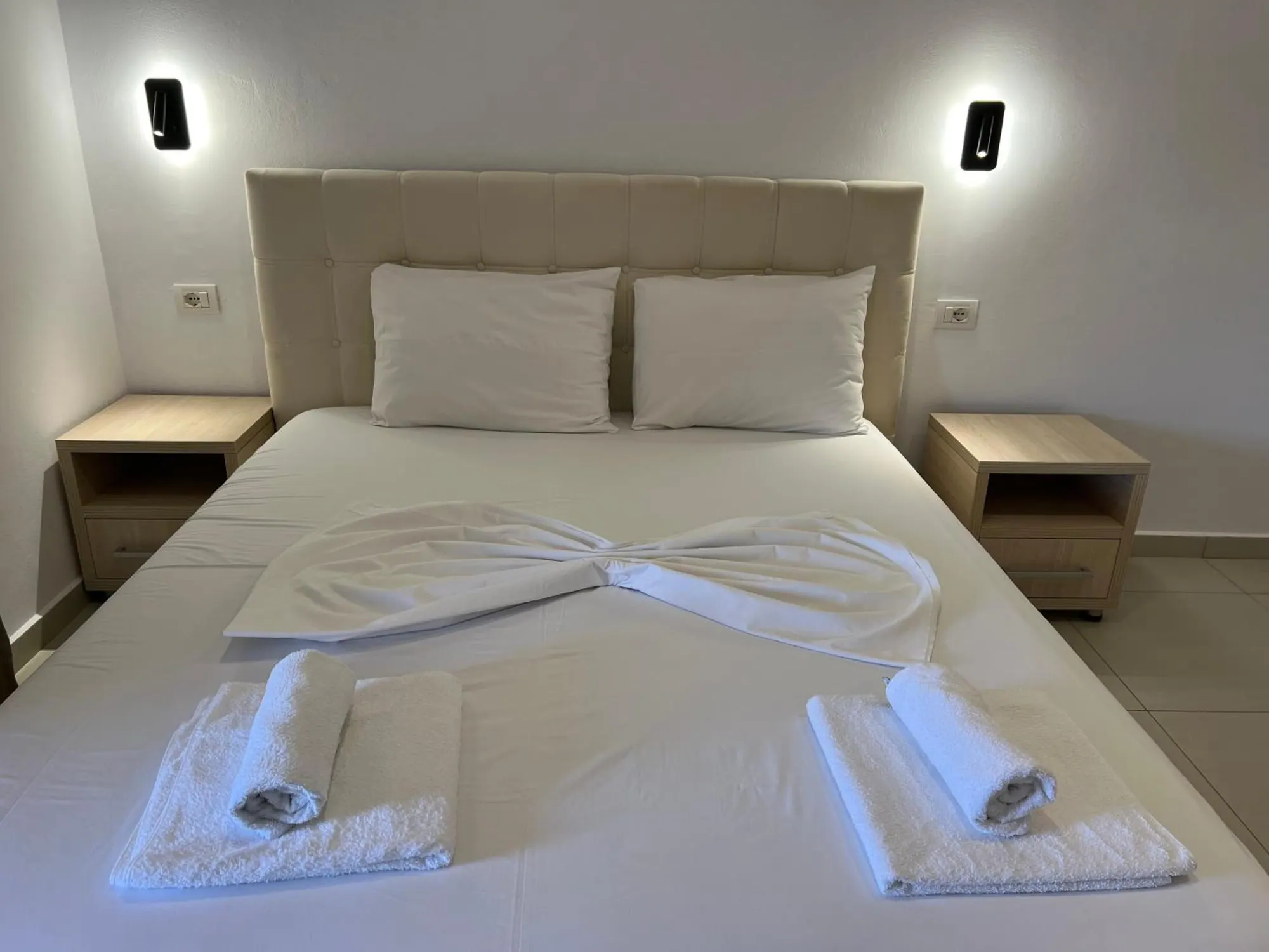 A large double bed with a linen headboard, oak bedside tables and two wall lights, towels folded on the white cover
